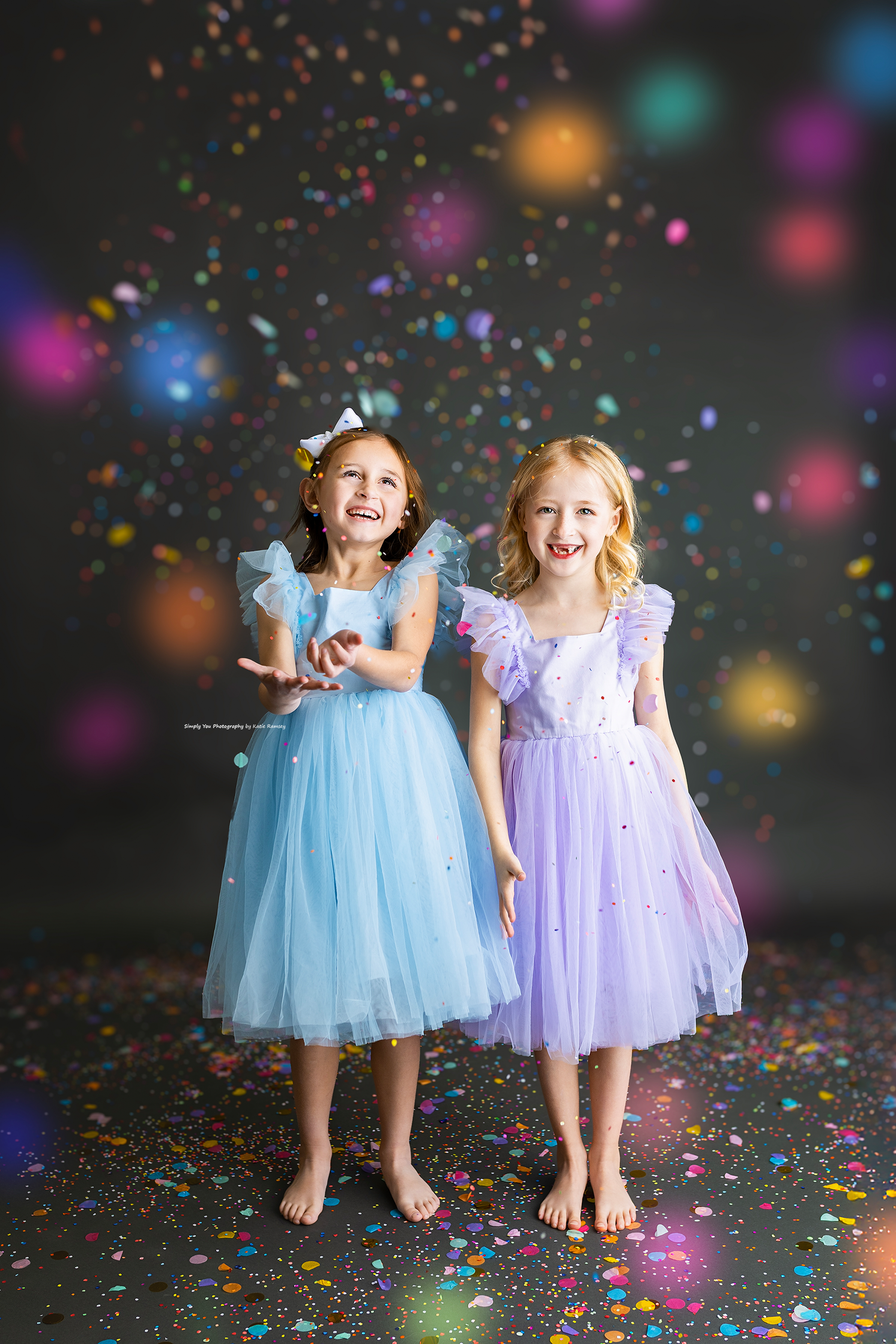 Two happy girls in blue and purple princess dresses laughing with colorful confetti falling.