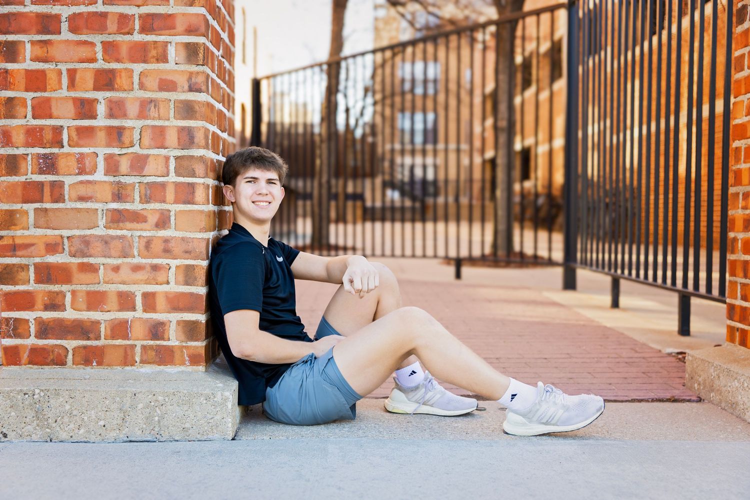 Teen boy in navy polo sitting against brick wall outdoors smiling.