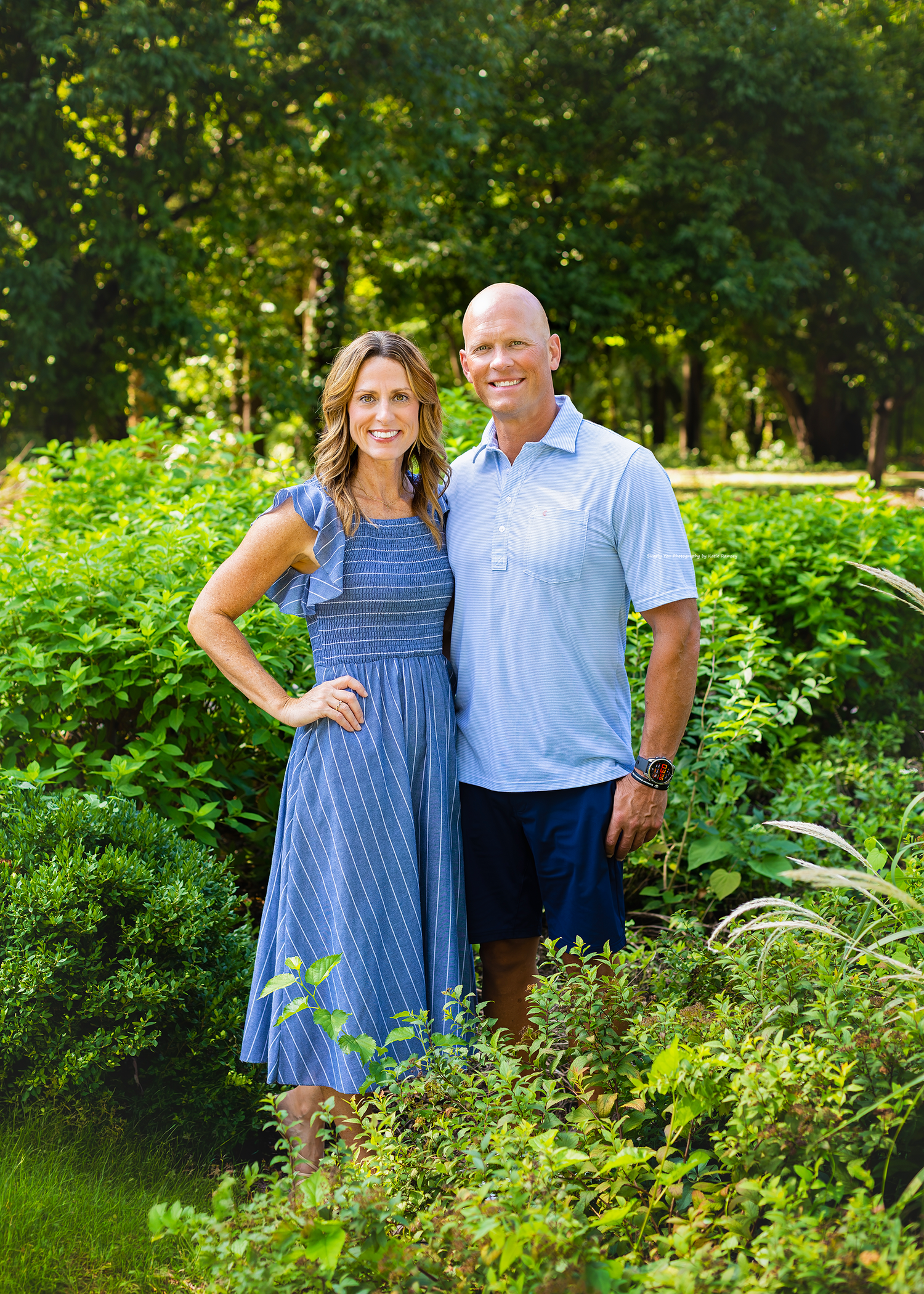 A smiling couple poses outdoors in a lush green garden, wearing coordinating blue outfits on a sunny day.