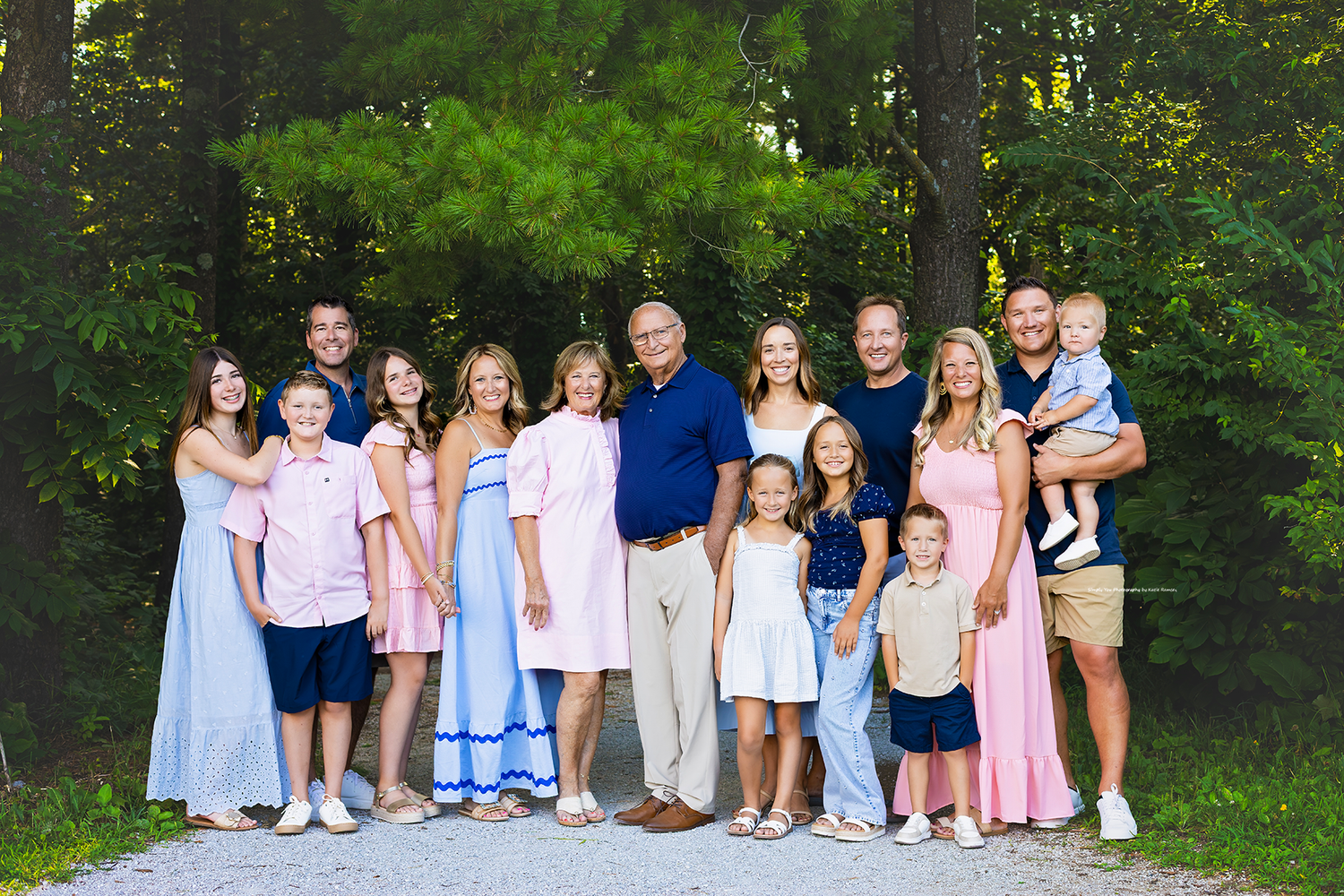 Large family group portrait smiling outdoors on a gravel path surrounded by lush green trees on a sunny day.