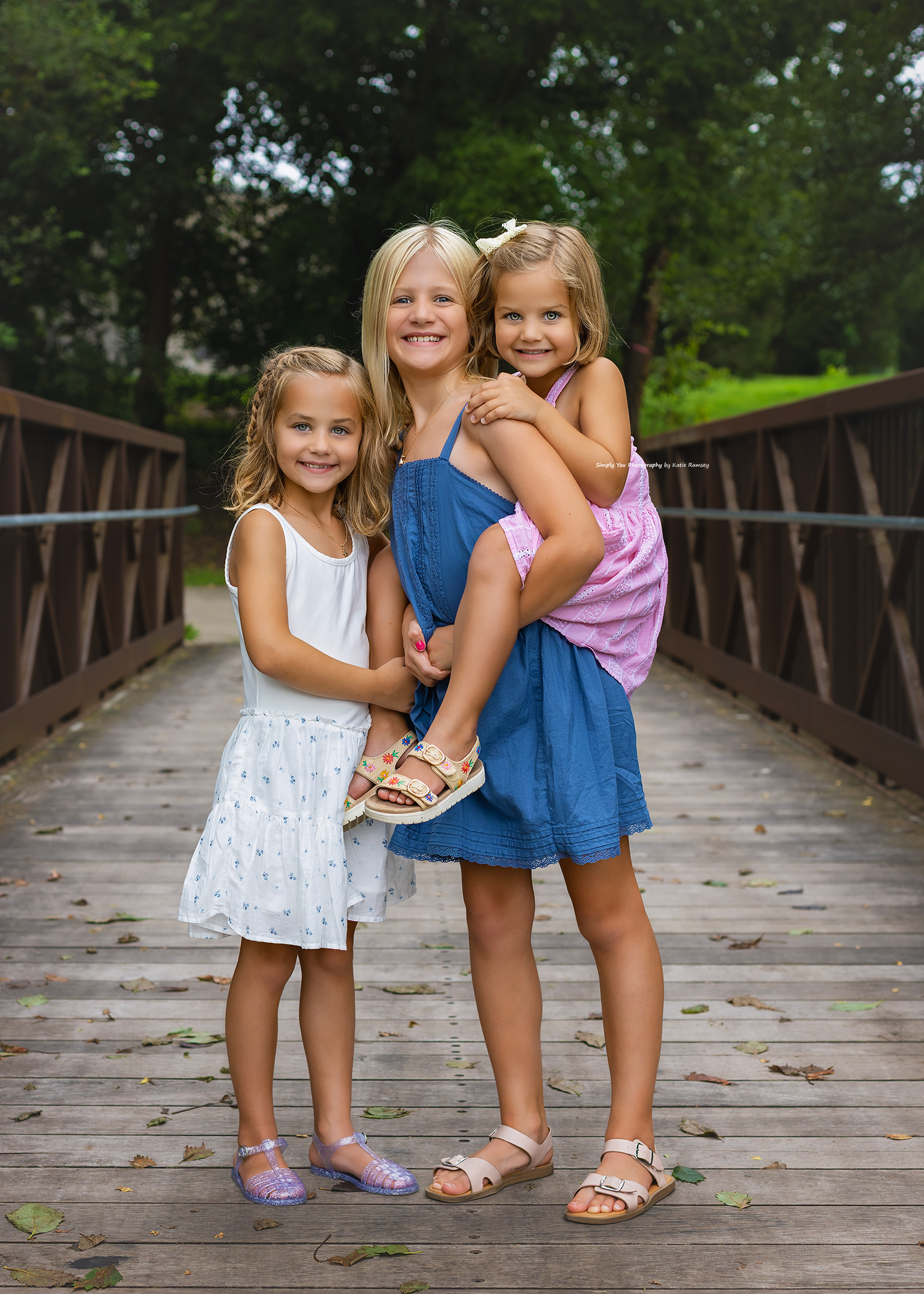 Three young girls pose together on a wooden bridge, smiling, wearing summer dresses and sandals outdoors.