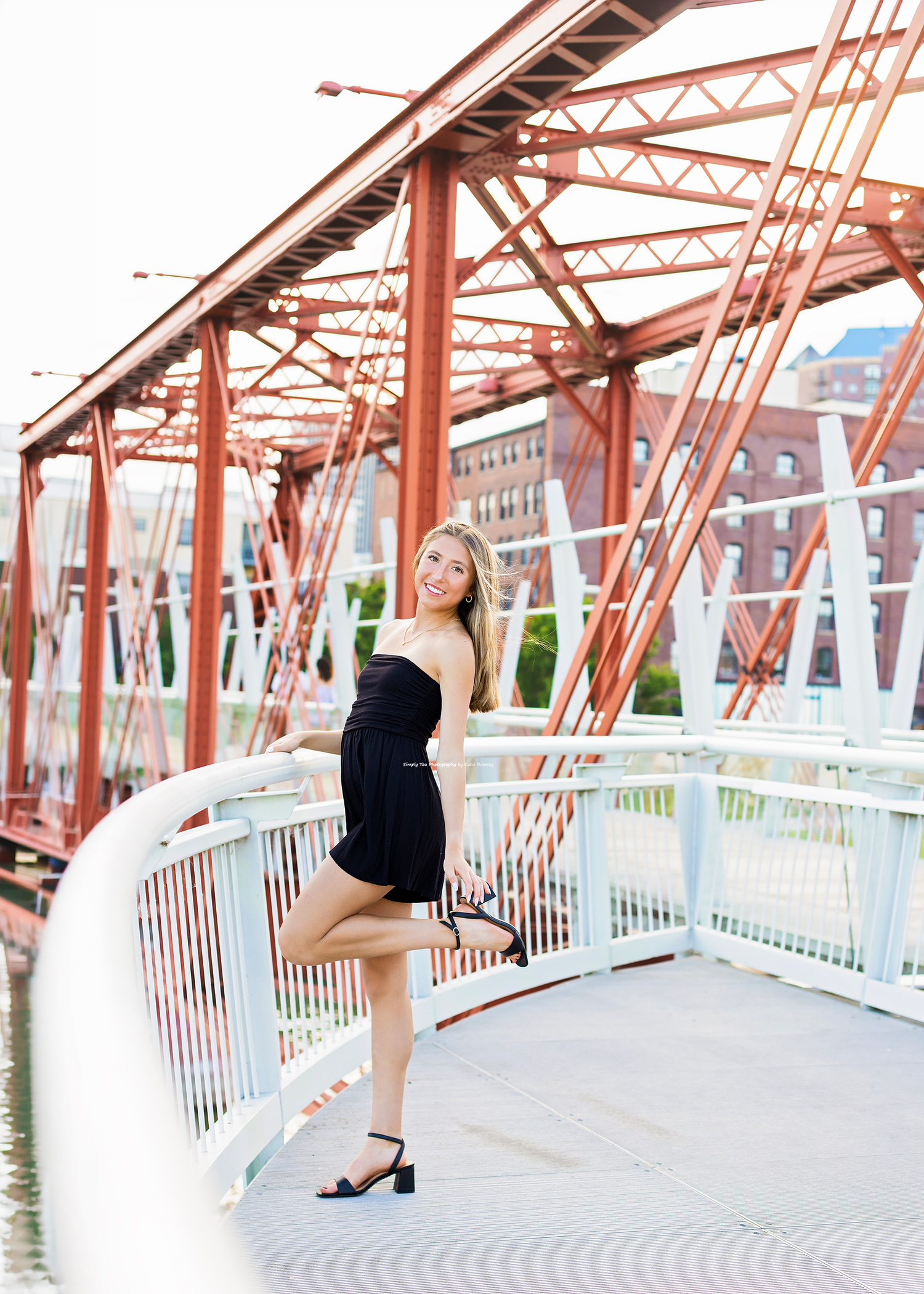 Young woman in black strapless romper posing on a red steel bridge with white railings and urban background.