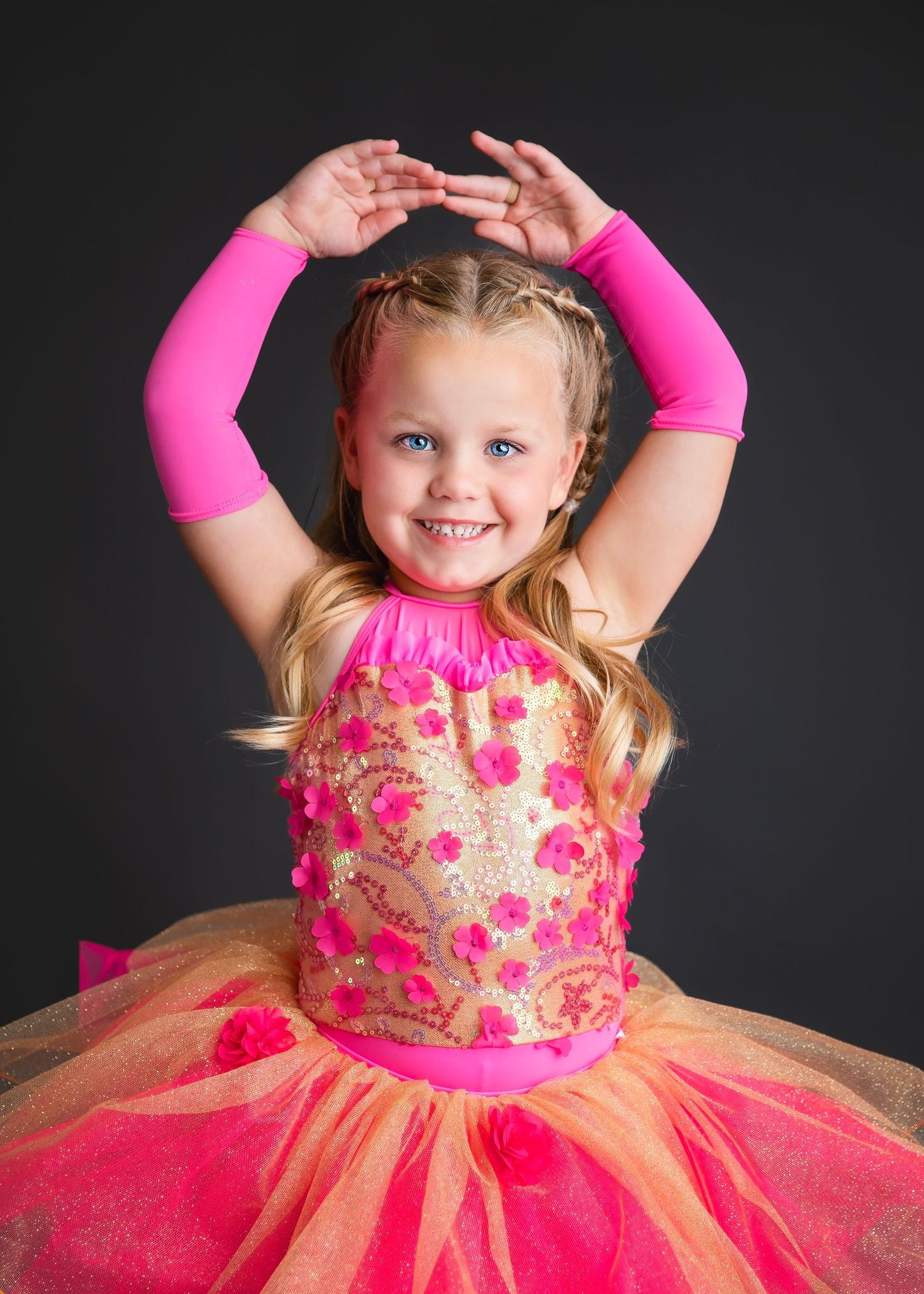 Young girl in pink and orange tutu dance costume posing with arms raised overhead in studio.