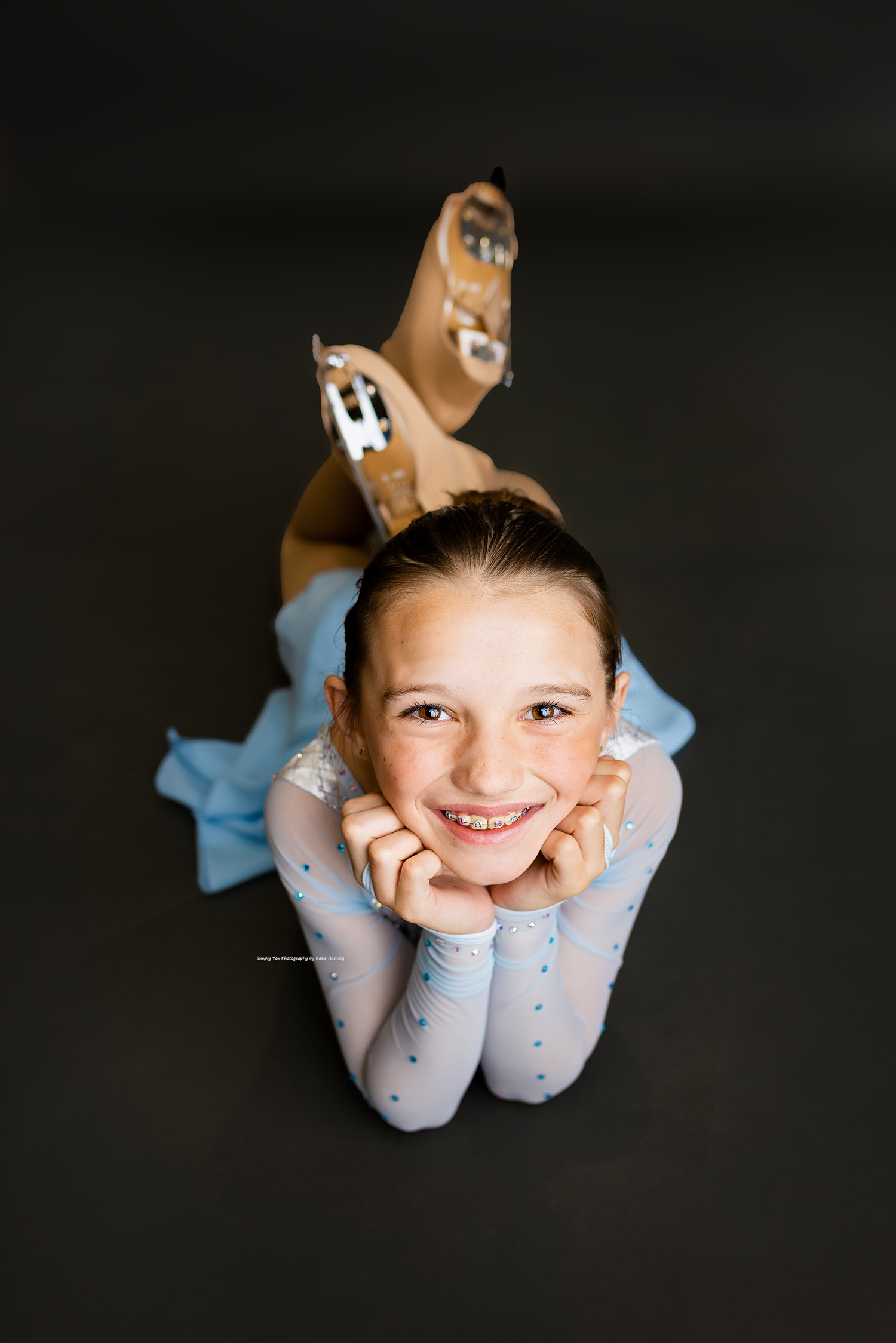 Young girl in light blue leotard lying on floor smiling, wearing ice skates, viewed from above.