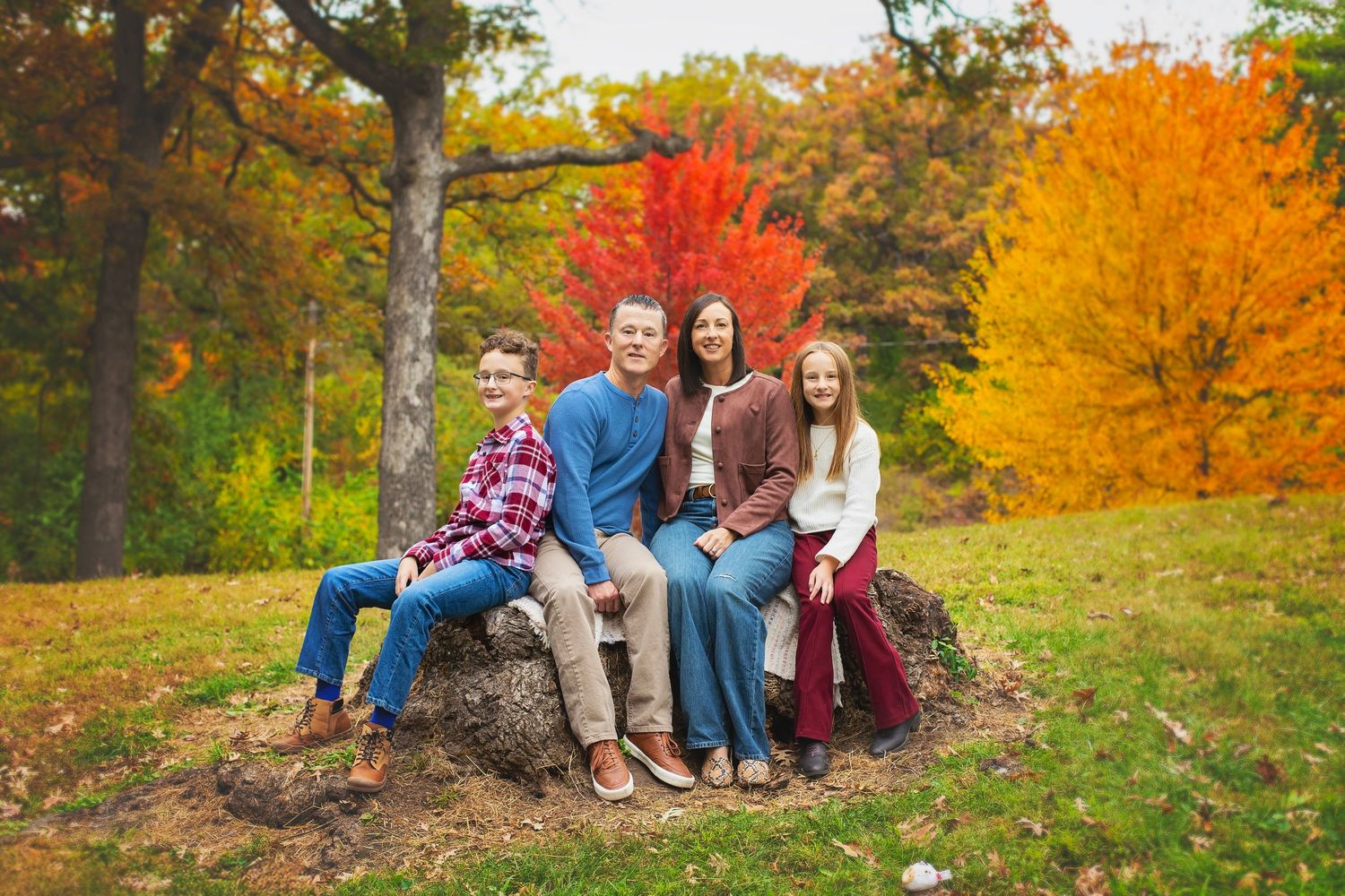 Family of four sitting on a large rock surrounded by colorful autumn foliage.
