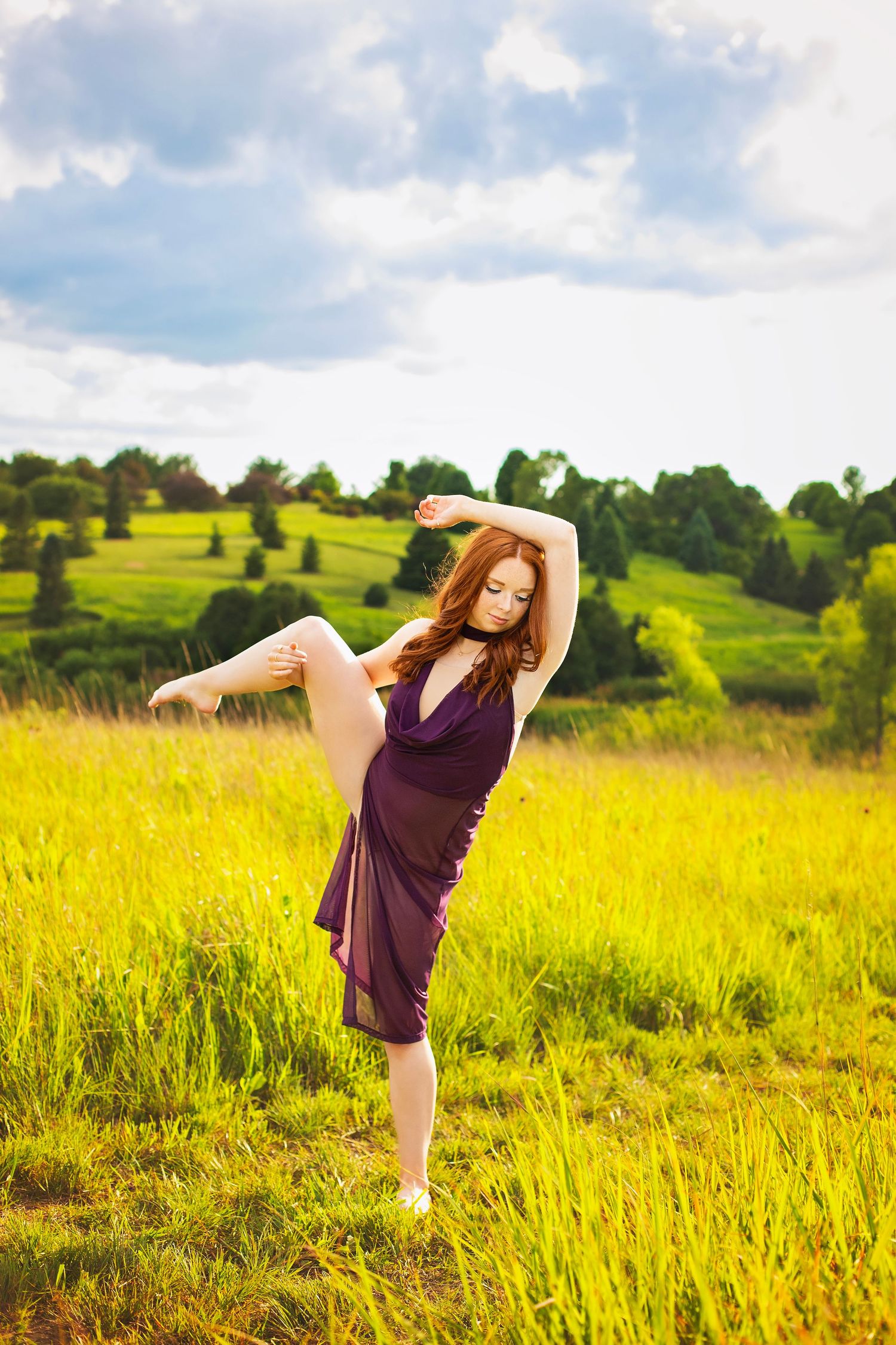 Graceful redhead dancer in purple dress performs arabesque pose in lush green field under cloudy sky.