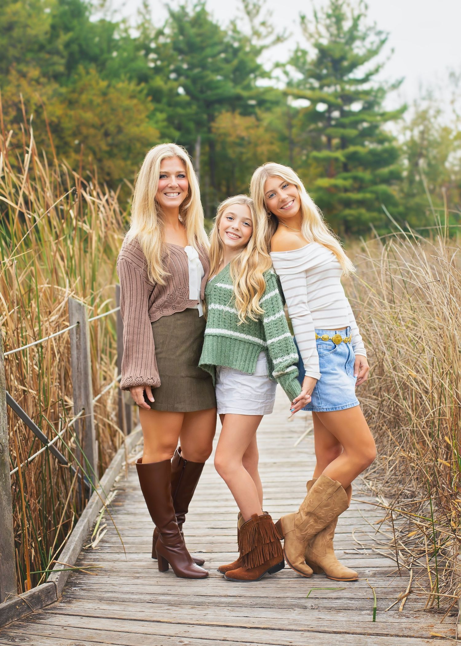 Three blonde girls pose together on a wooden boardwalk surrounded by tall golden autumn grasses.