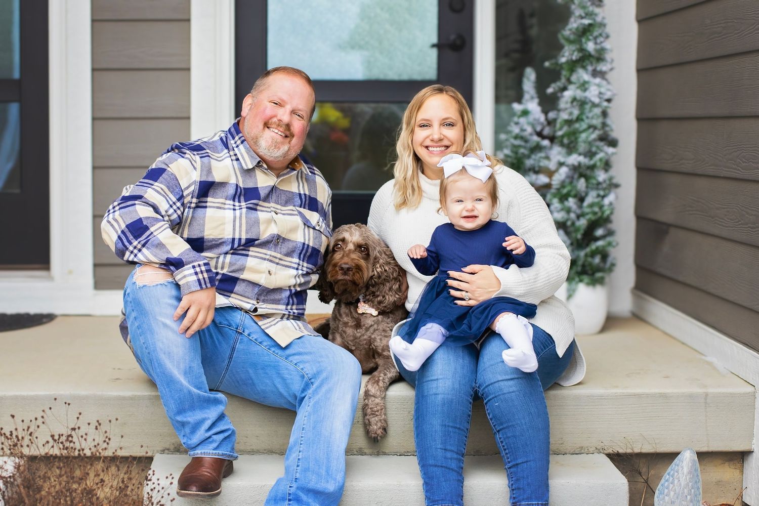 Happy family with baby girl and brown dog sitting on front porch steps with winter decorations.