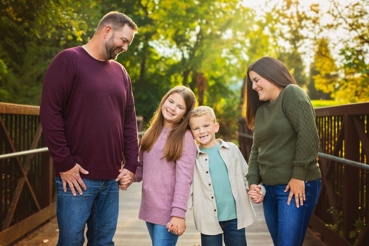 A cheerful family of four holds hands on a sunlit wooden bridge in autumn.