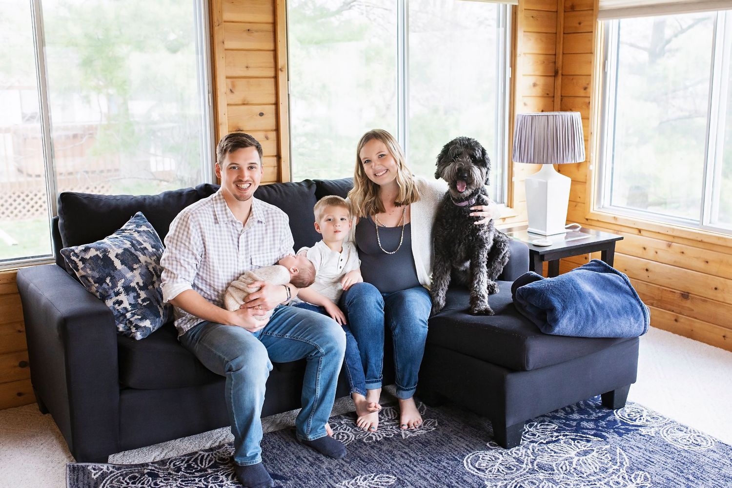 Family of three with a large black dog sitting together on a sofa in a wood-paneled room.