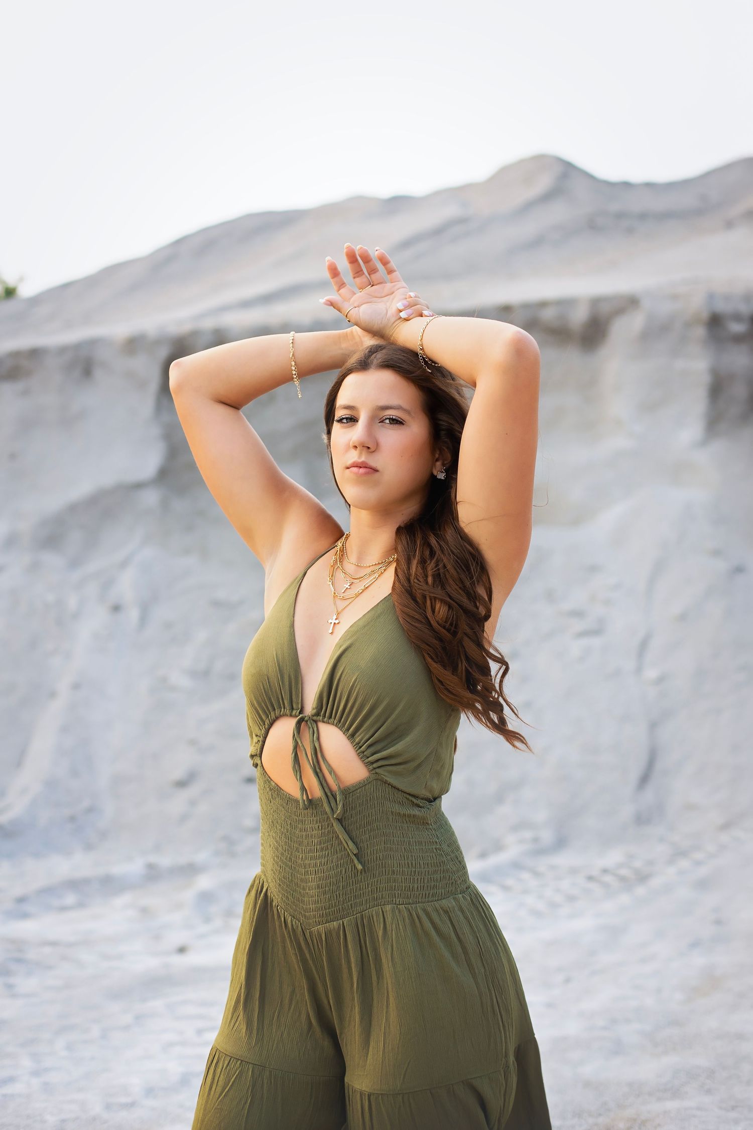 Young woman in olive green jumpsuit poses with arms raised before a sandy quarry backdrop.