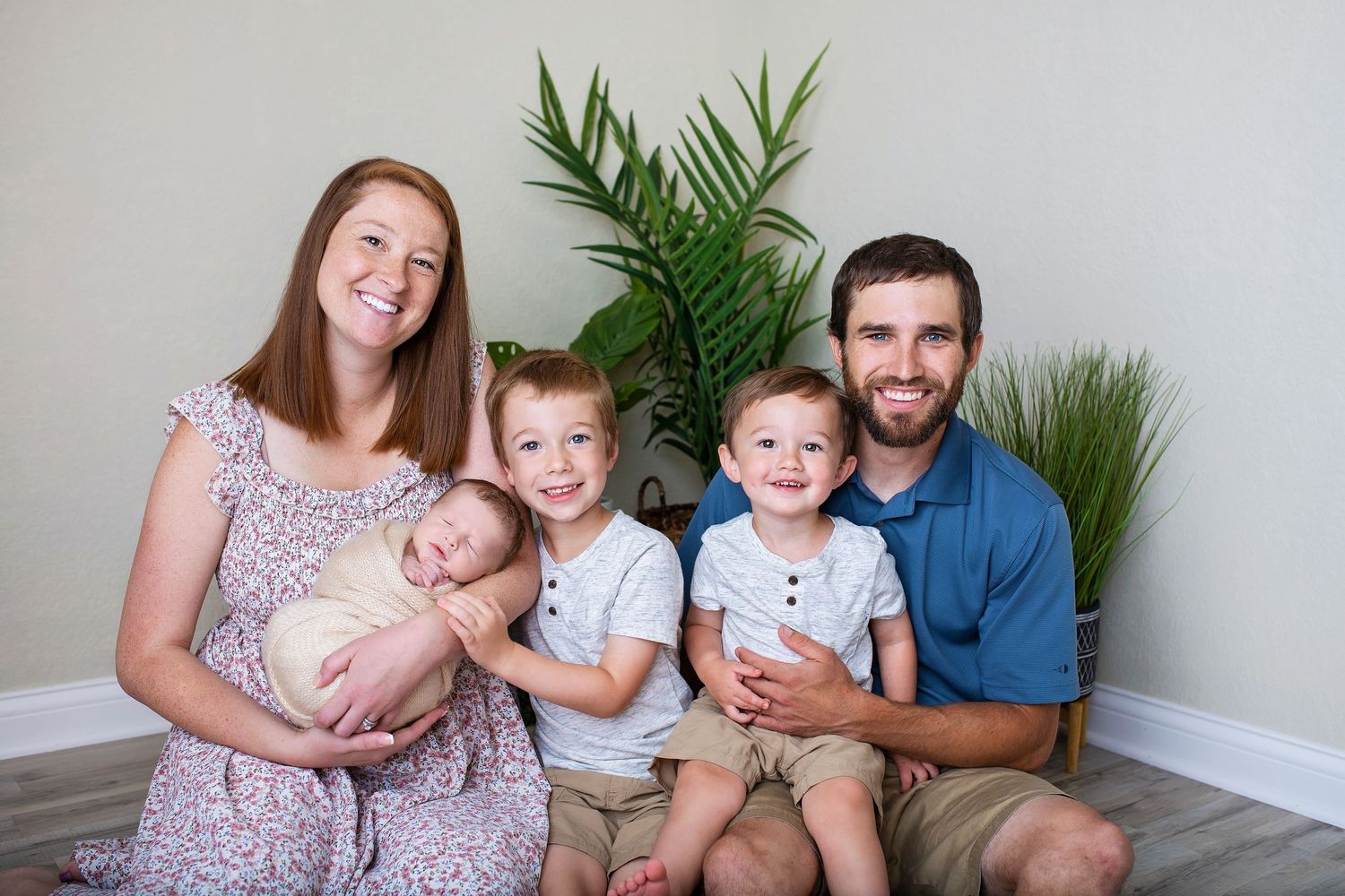 Smiling family portrait with parents, newborn baby, and two young boys in front of green plants.