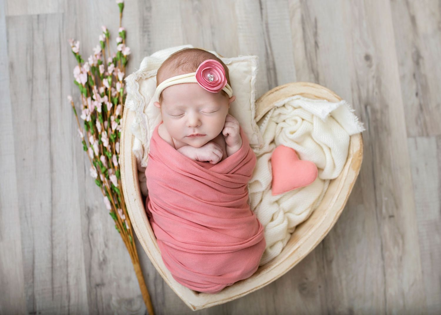 Newborn baby girl swaddled in pink wrap sleeping in heart-shaped wooden bowl with floral headband.