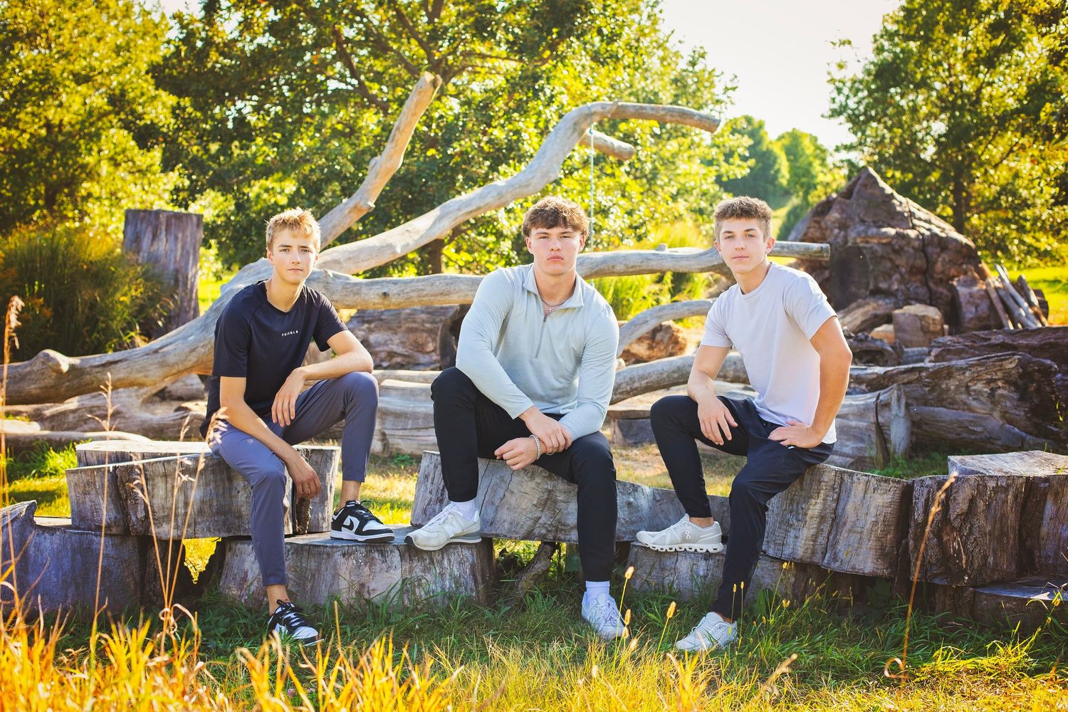 Three teenage boys sit on wooden logs outdoors in golden sunlight for senior portraits.