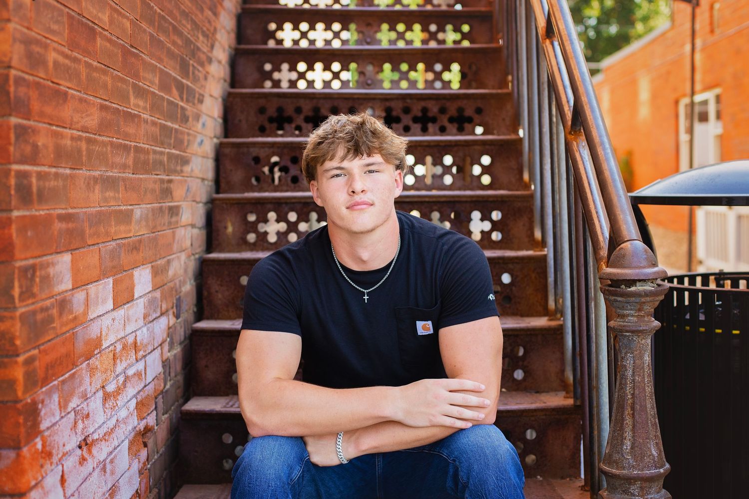 Teen boy in black t-shirt sits on brick stairs with arms crossed for senior photos.