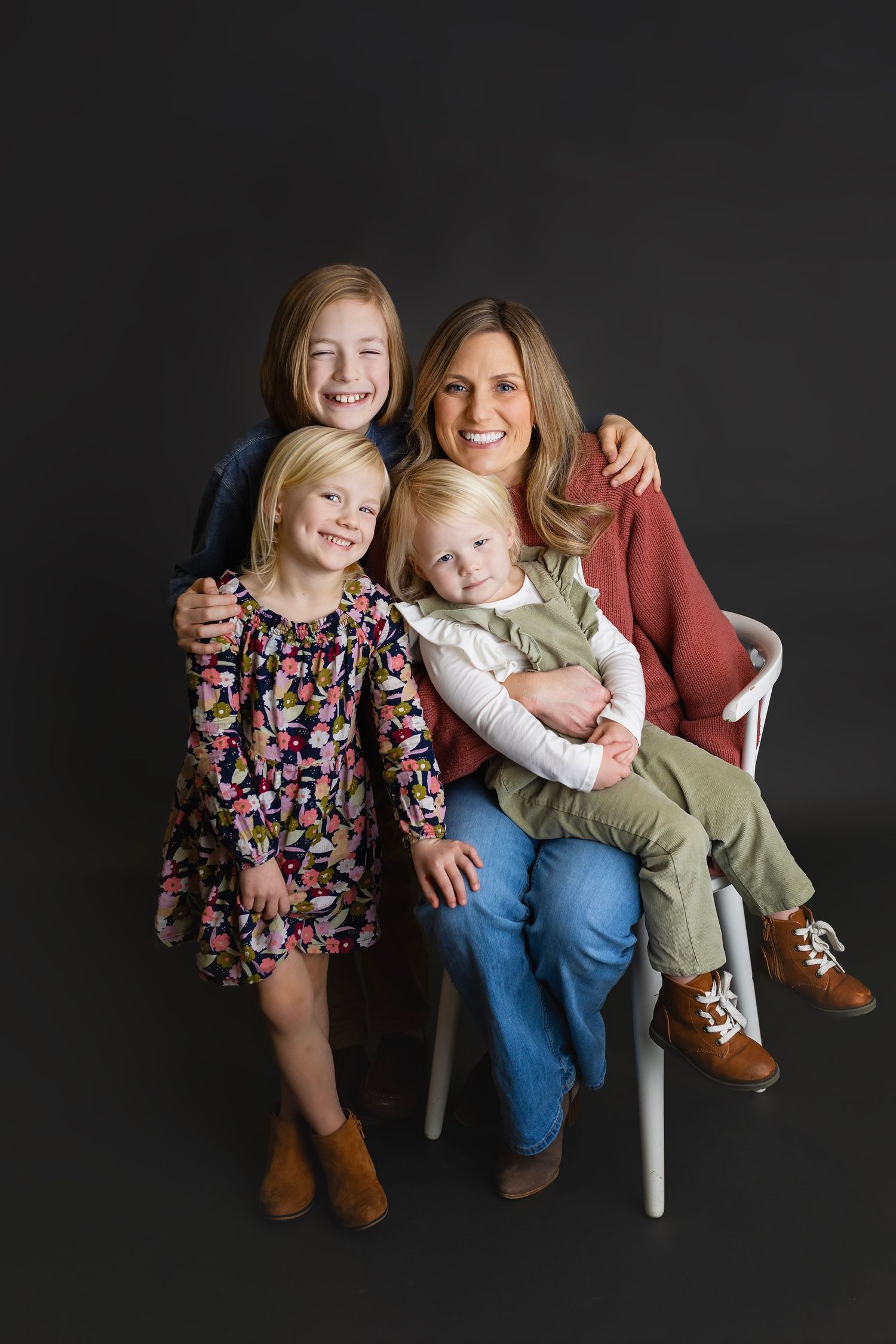 Smiling mother seated on white chair with three blonde daughters hugging her in studio family portrait.