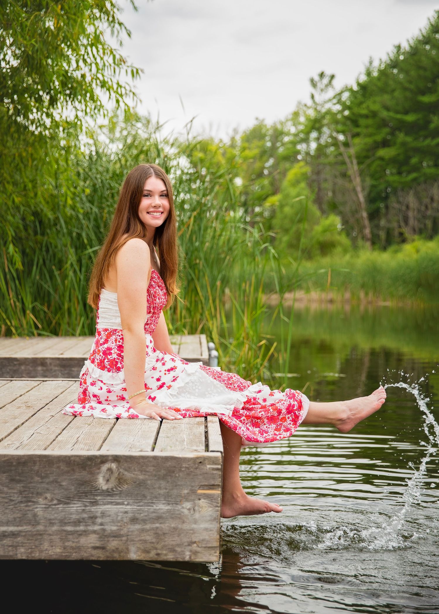 Young woman in red floral dress sitting on wooden dock splashing feet in calm lake surrounded by lush greenery.