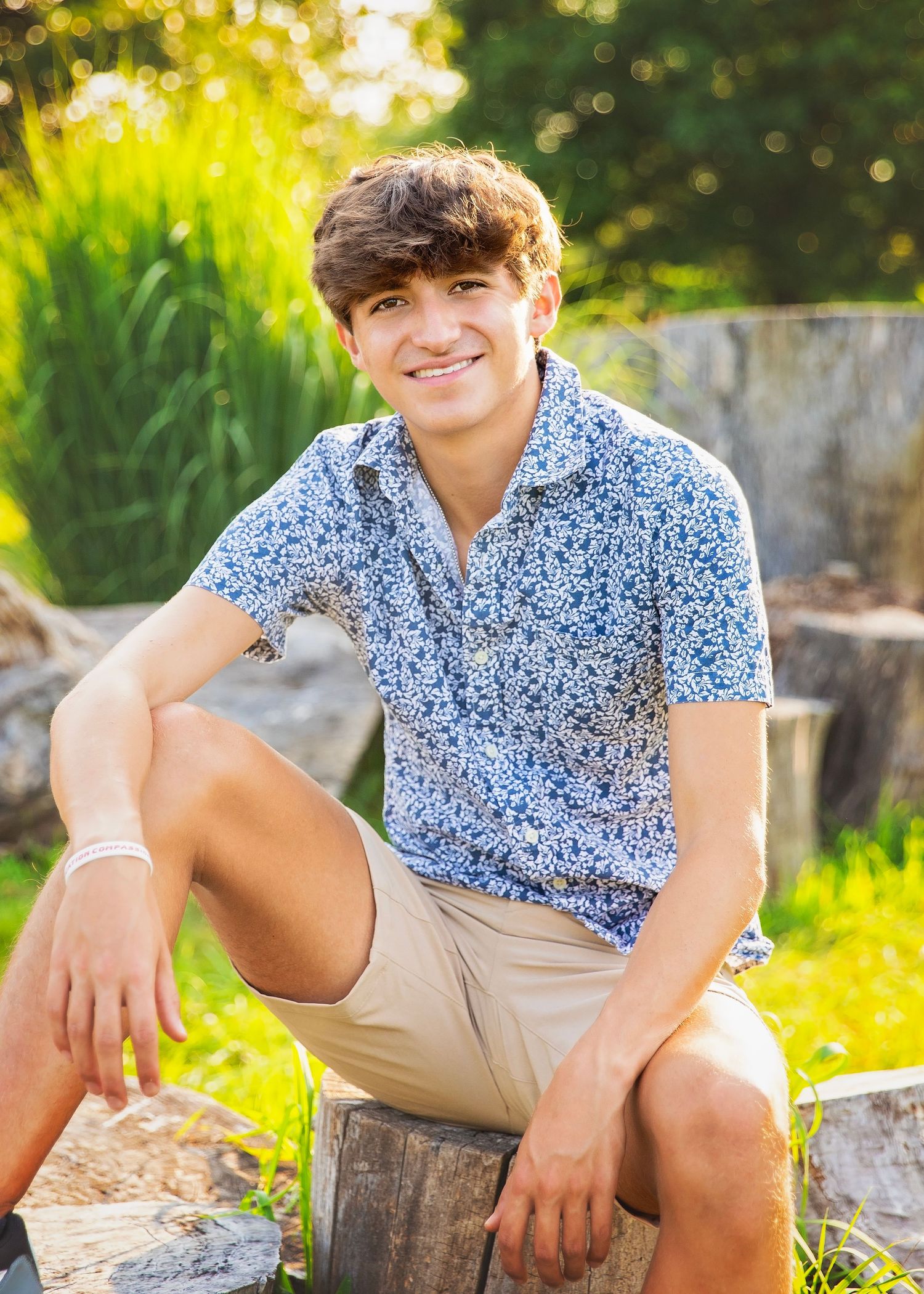 Smiling teenage boy in blue floral shirt and khaki shorts sitting on a wooden stump in a sunny garden.