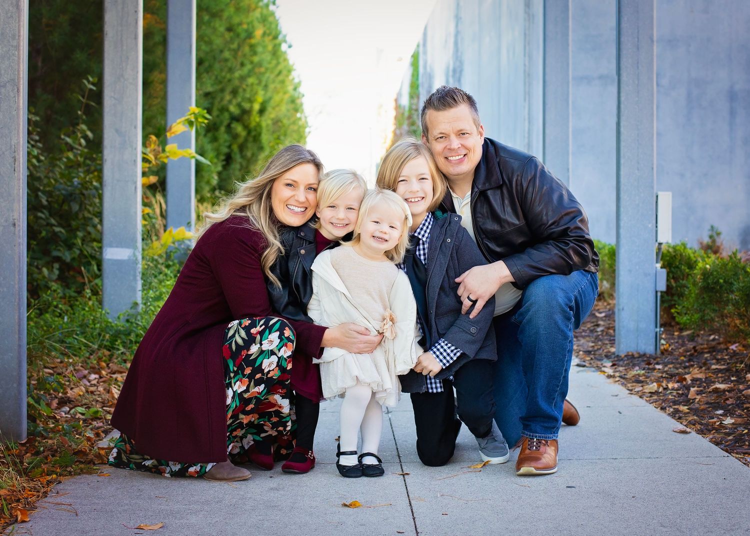 A cheerful family of five poses on a sidewalk outdoors, with two parents kneeling beside three young children.