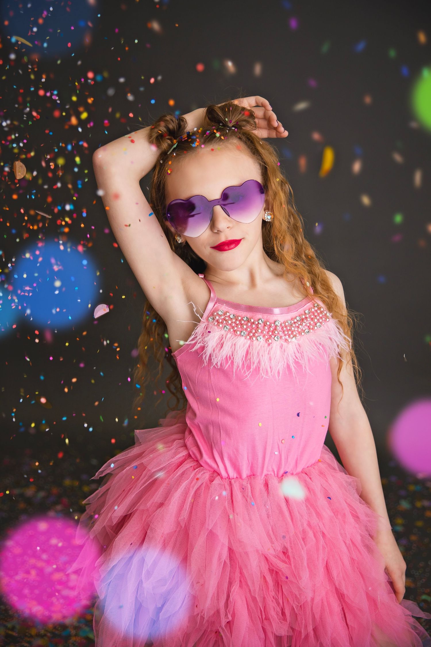 Girl in pink tutu dress posing with purple heart sunglasses amid falling colorful confetti on dark background.