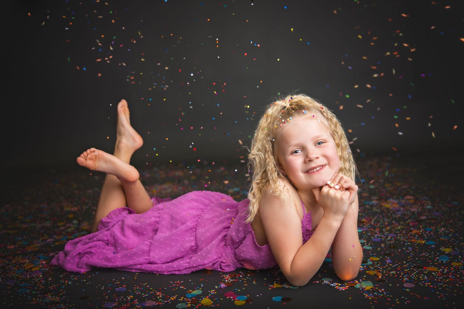 Smiling blonde girl in pink dress lying on floor with colorful confetti on dark background.