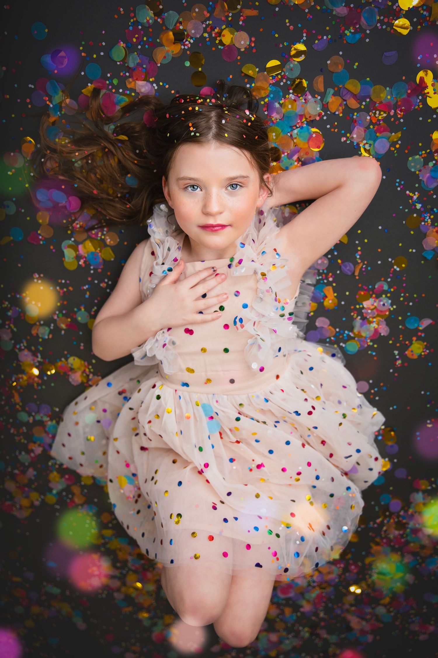 Young girl in polka dot dress lying among colorful confetti, posing for a festive photo shoot.