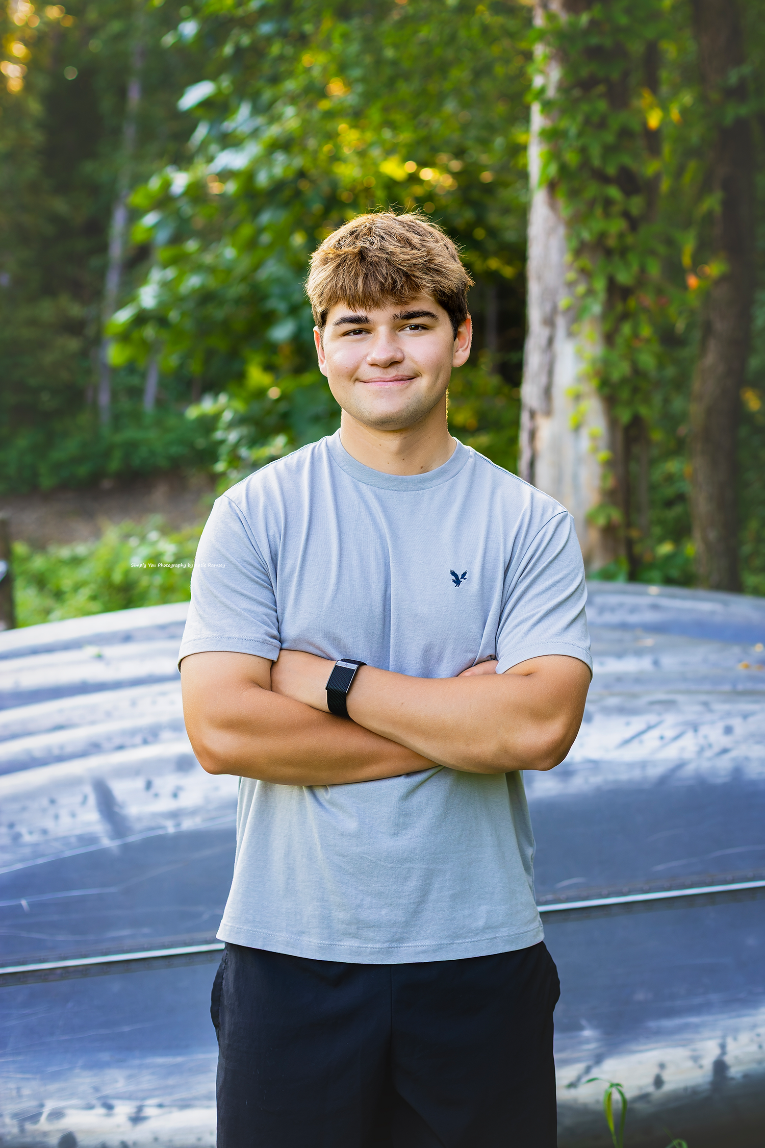 Young man in gray American Eagle t-shirt stands with arms crossed, smiling outdoors near trees.