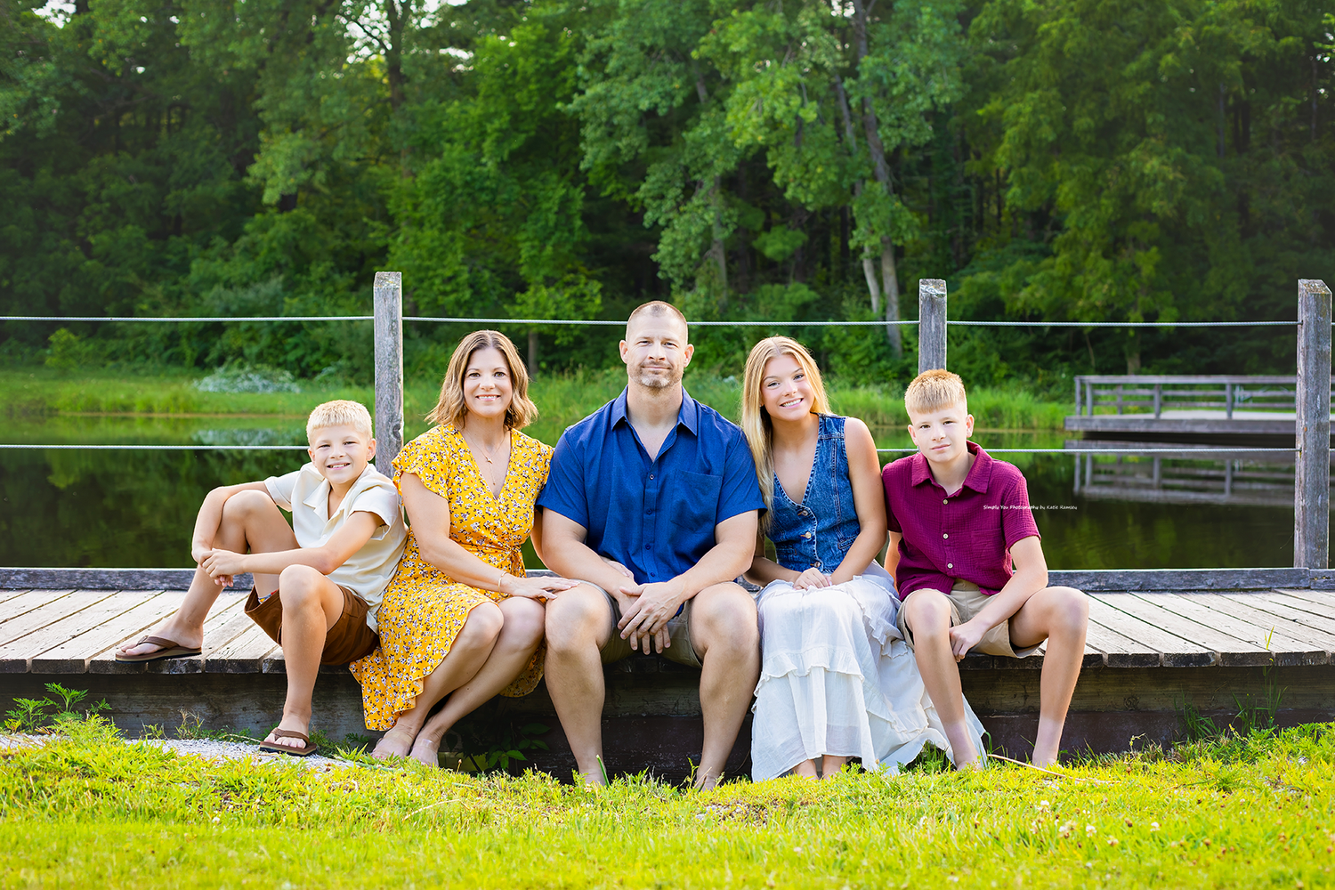 Family of five smiling together on a wooden dock surrounded by lush green trees near a serene lake.