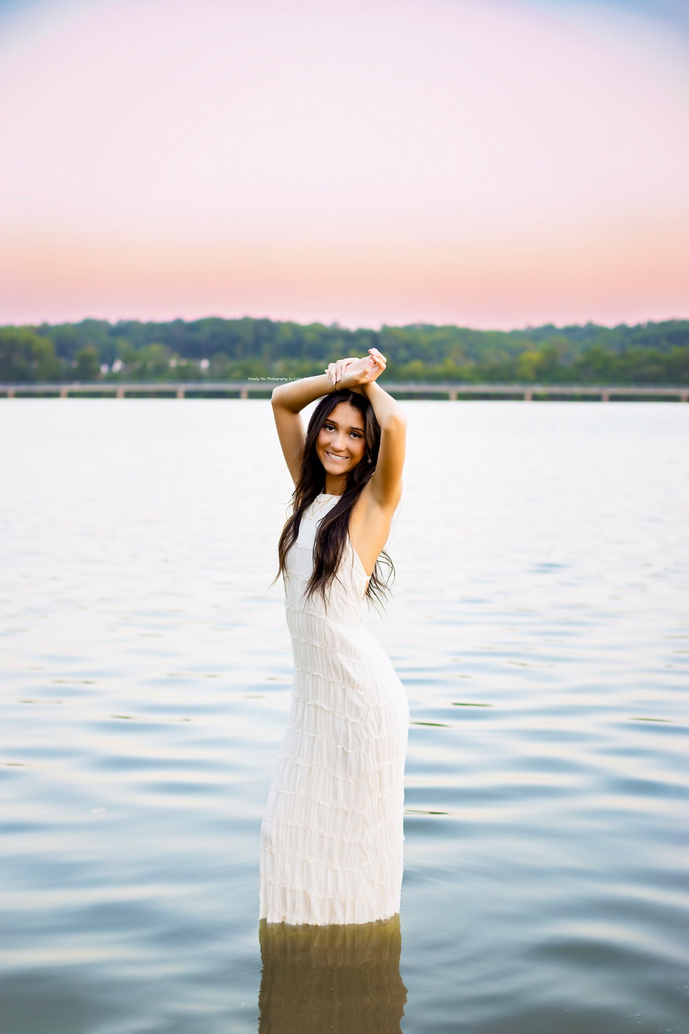 Young woman in white dress standing in a lake at dusk with arms raised above her head.