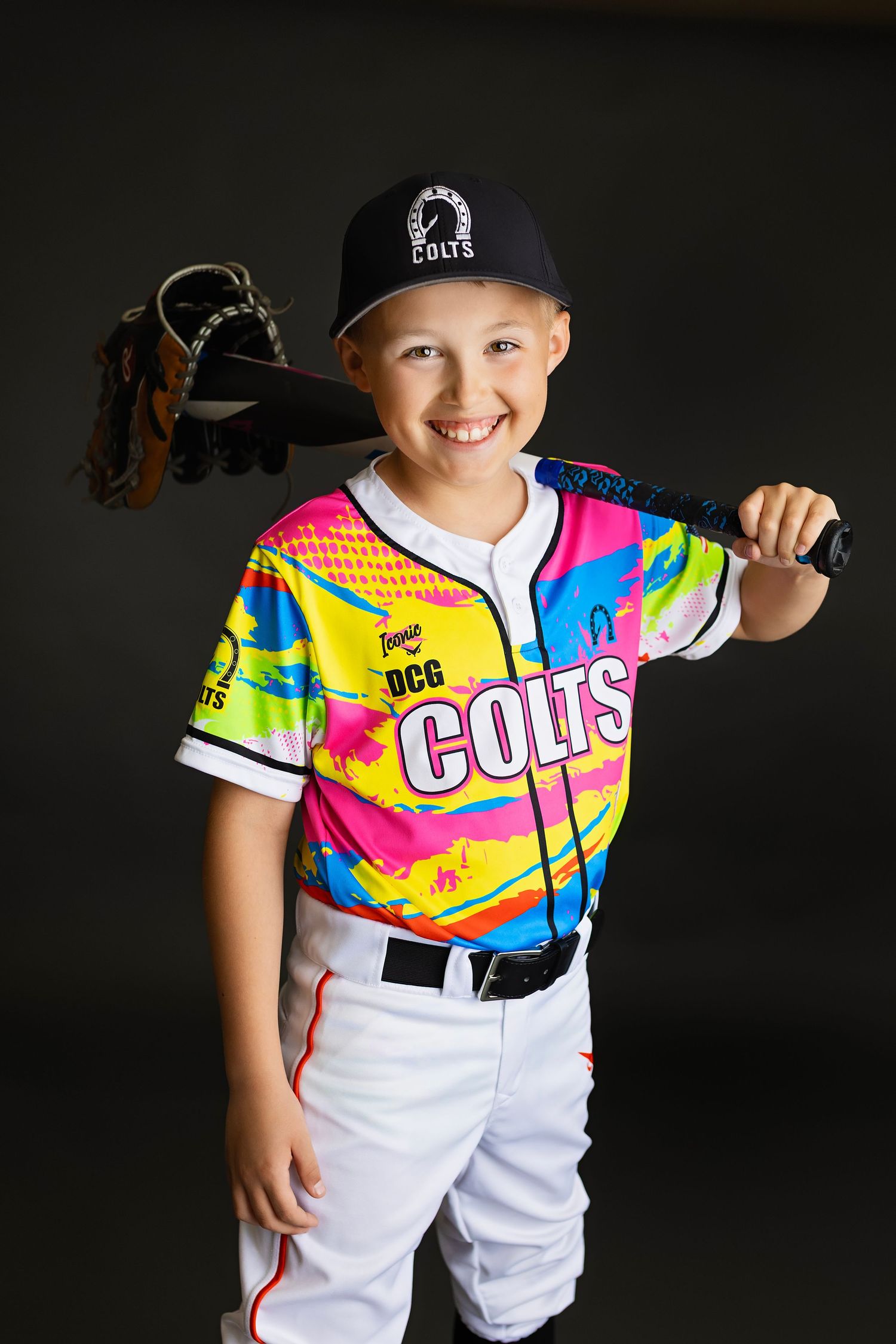 Smiling young boy in colorful Colts baseball jersey holding bat and glove over shoulder on dark background.