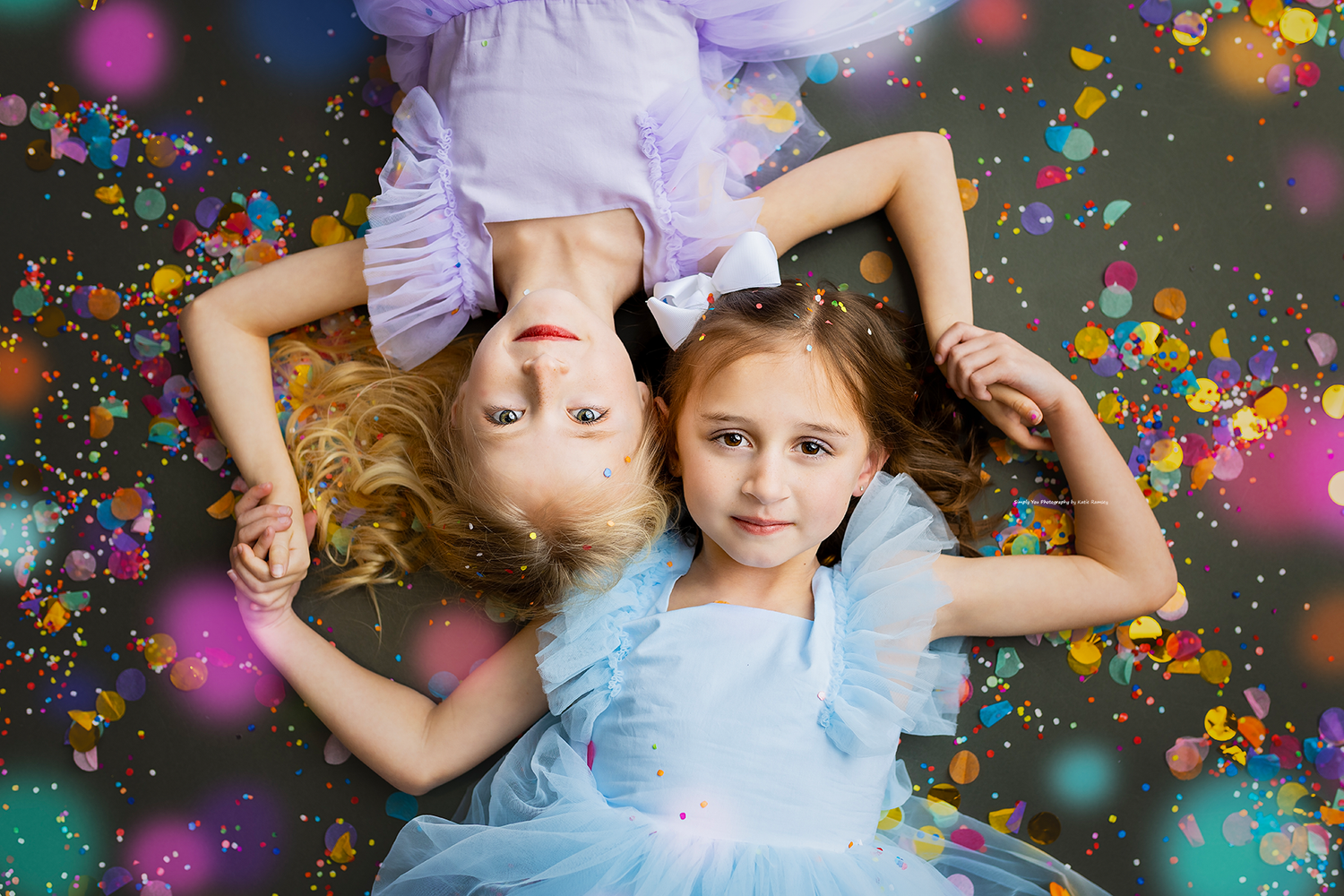 Two young girls in blue and purple tulle dresses lying on floor surrounded by confetti.