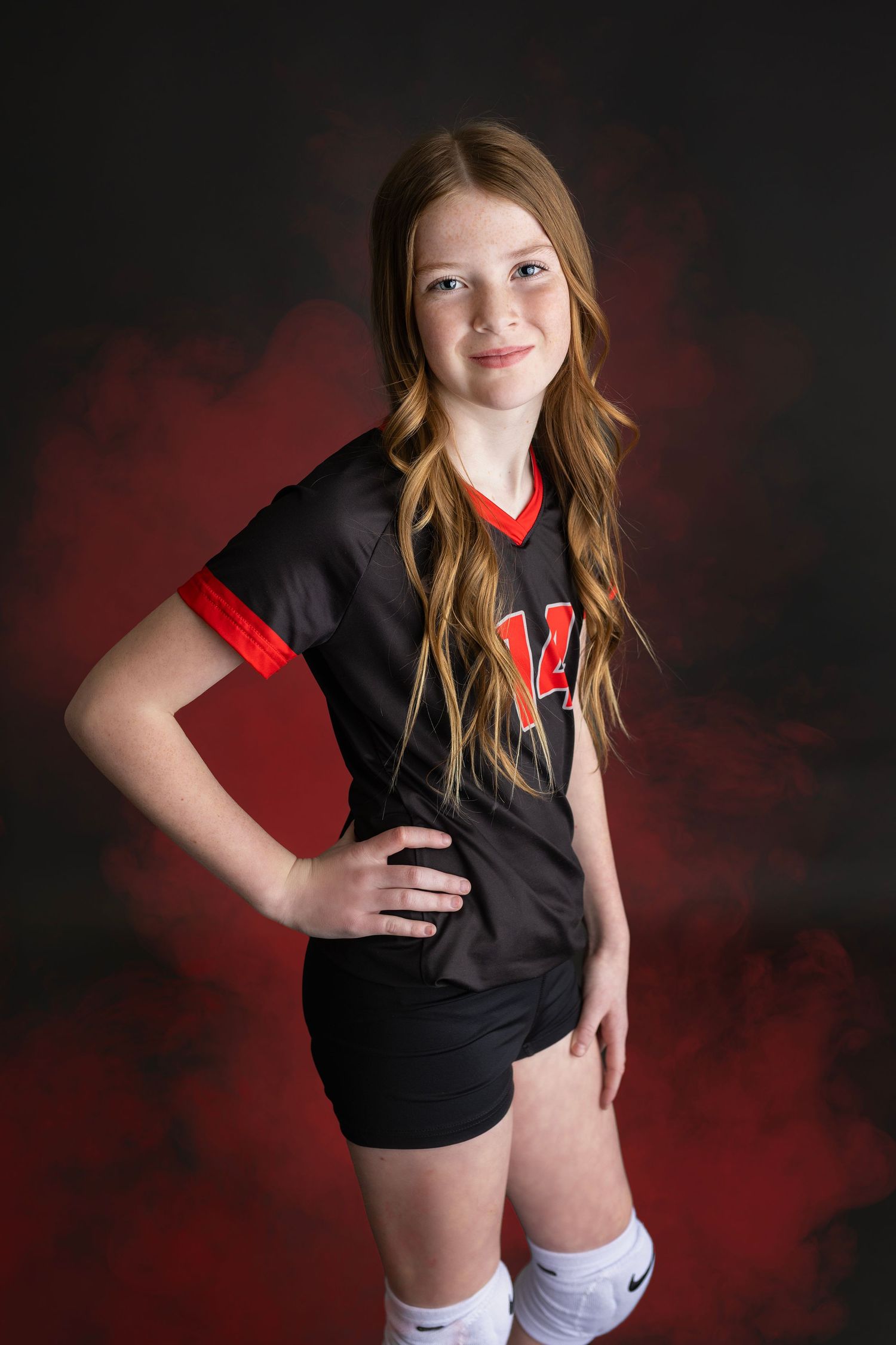 Young girl in black number 14 volleyball uniform posing with hand on hip against smoky red backdrop.