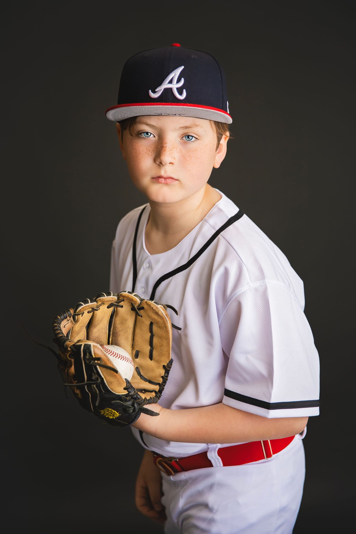 Young baseball player in Atlanta Braves uniform holding glove with ball, serious expression, dark background.