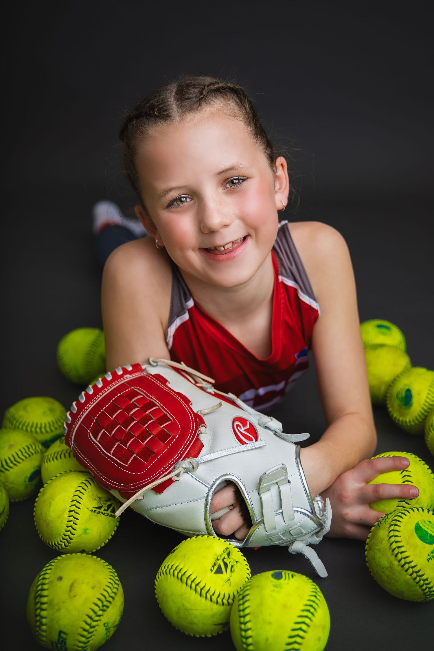 Smiling young softball player with Rawlings glove poses among yellow softballs in studio.