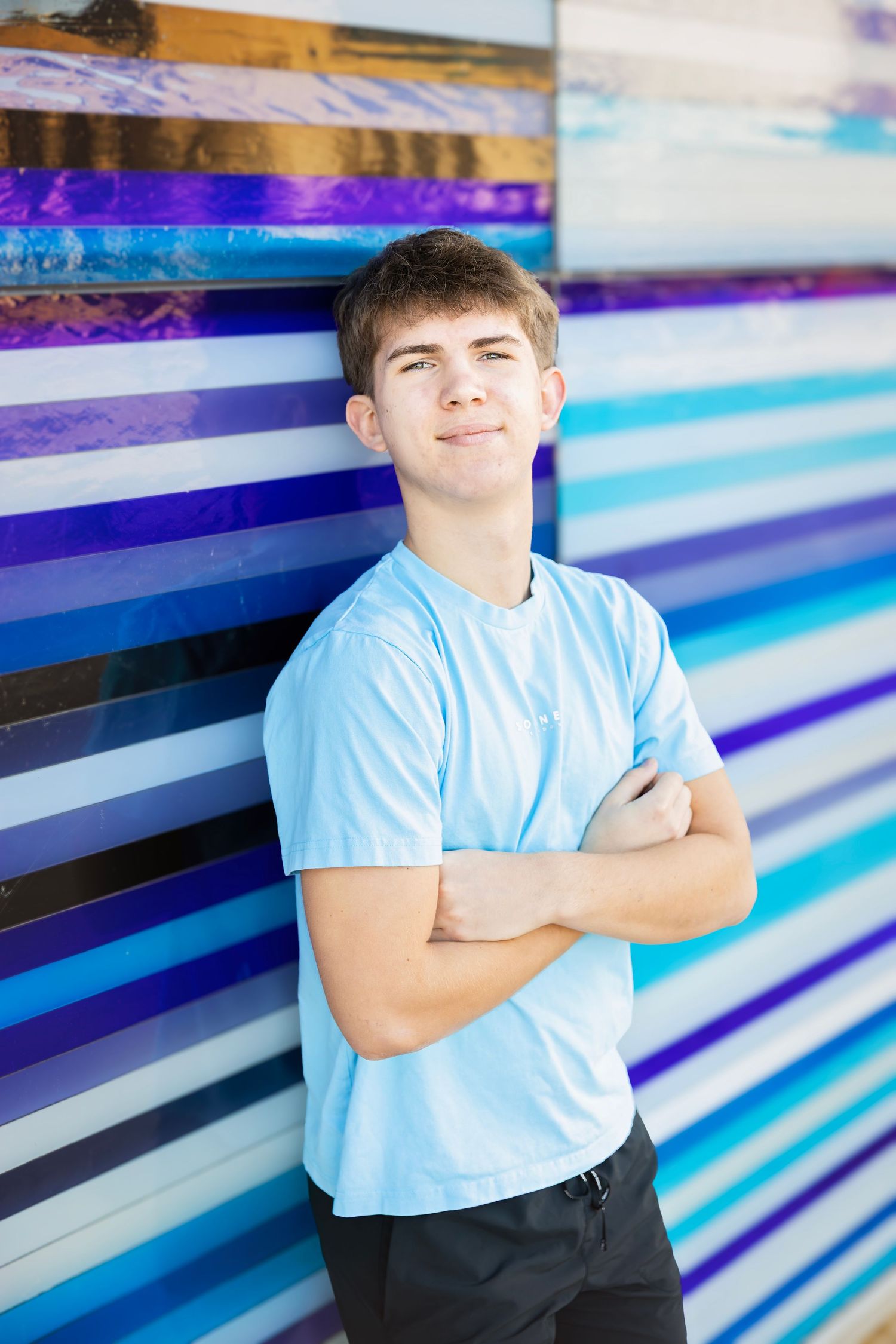 Teen boy in light blue shirt with arms crossed leaning against colorful striped wall.
