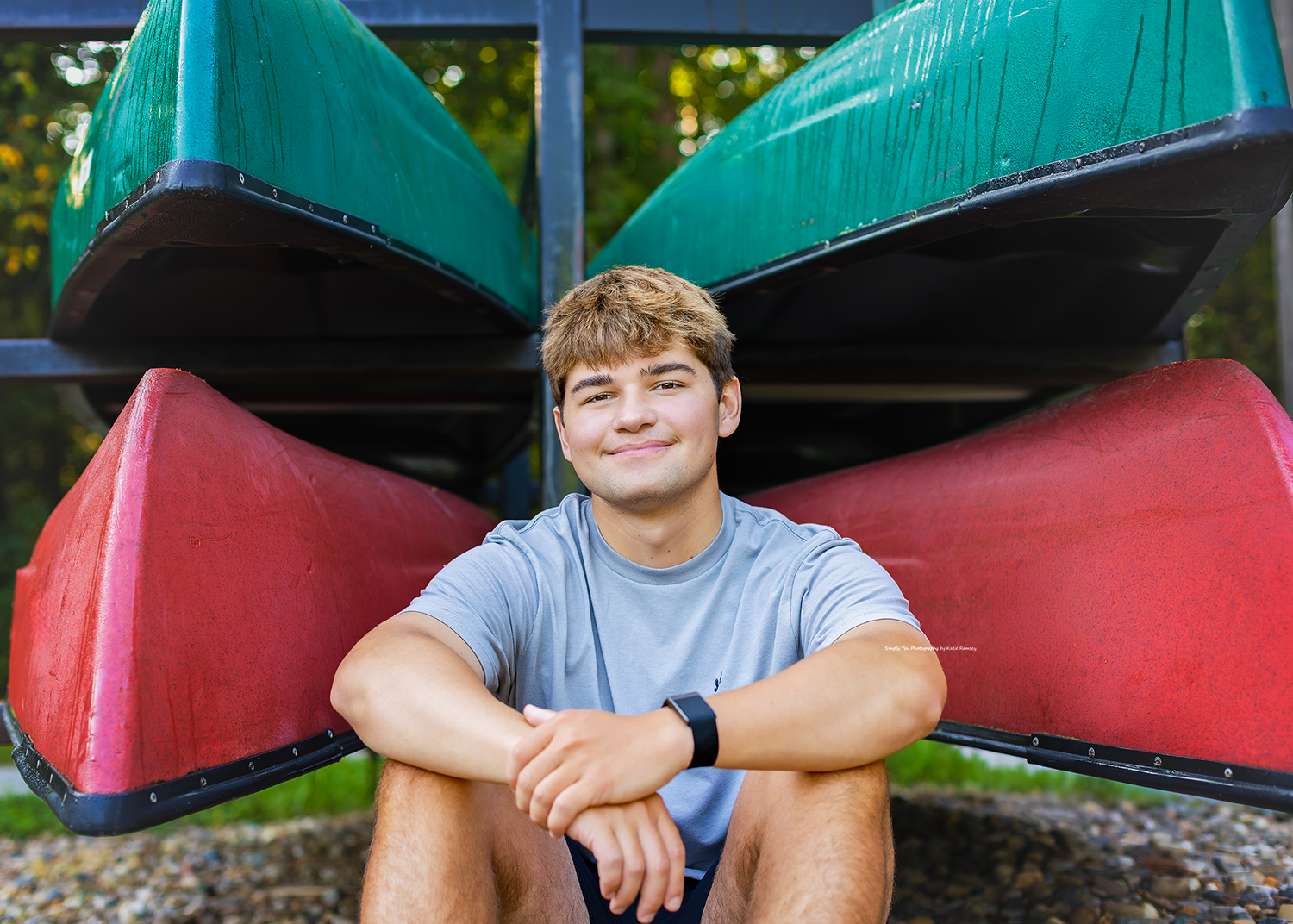 Smiling teenage boy sitting cross-legged in front of stacked red and green canoes outdoors.