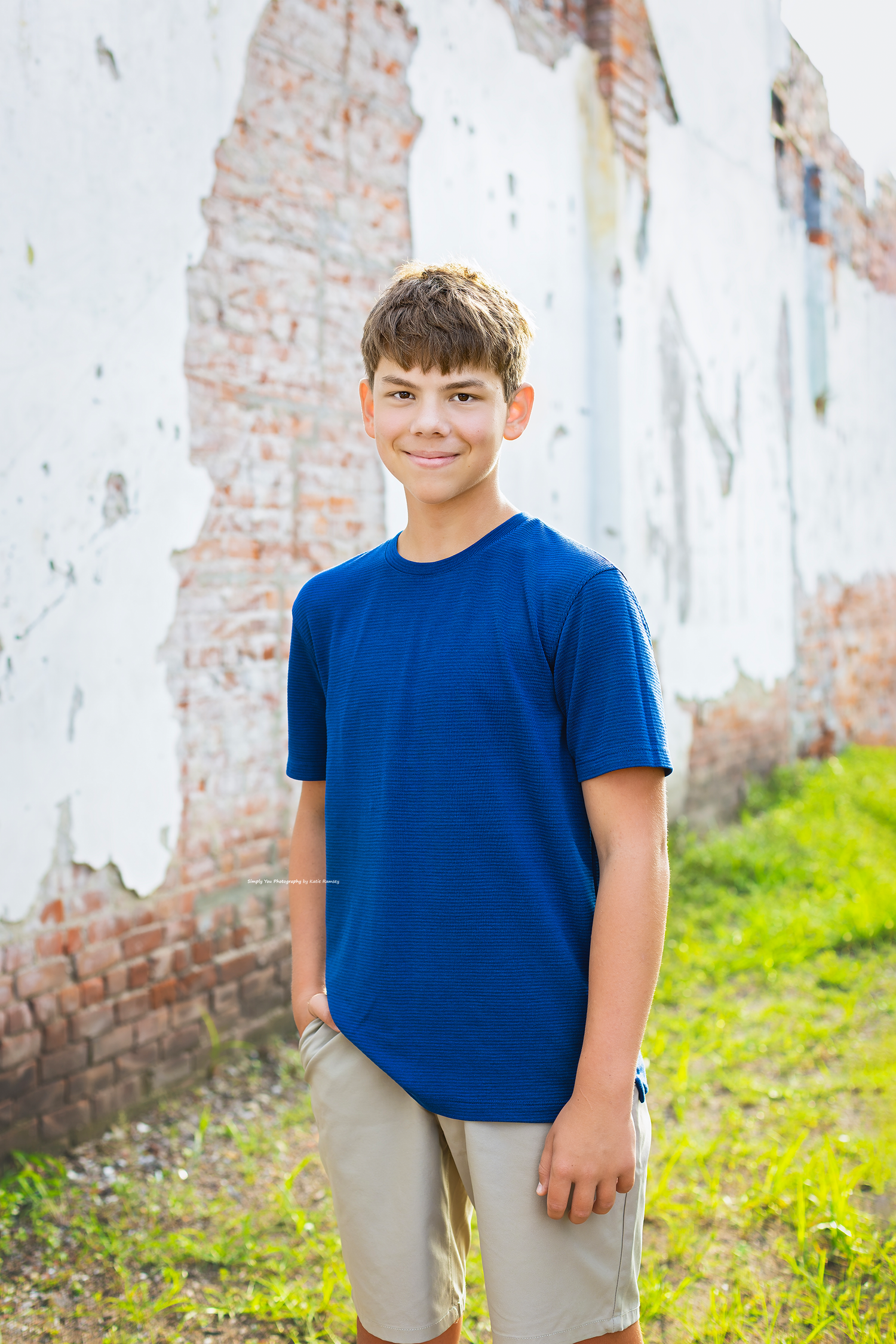 Smiling teenage boy in blue t-shirt and khaki shorts standing in front of a weathered brick wall.