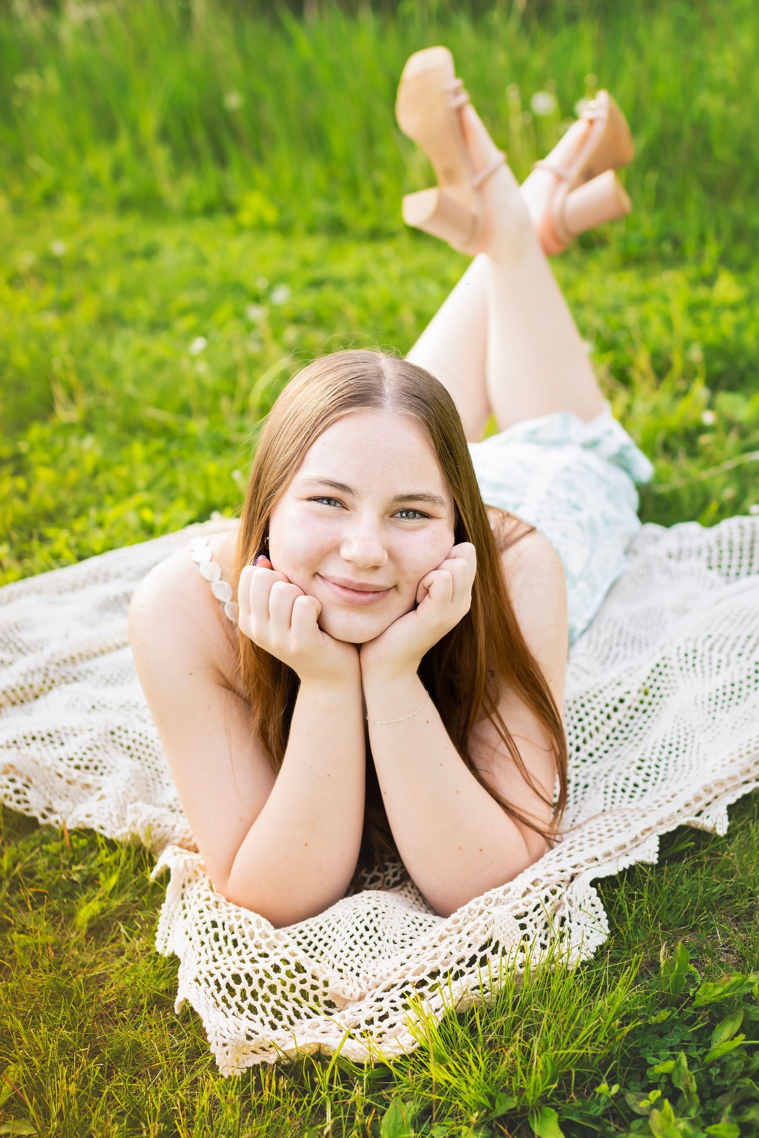 Smiling teenage girl lying on crochet blanket in green grass resting chin on hands with feet up.