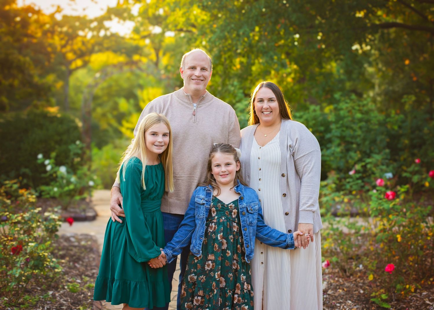 Family of four posing in a rose garden, parents standing with two daughters in green and floral dresses at sunset.