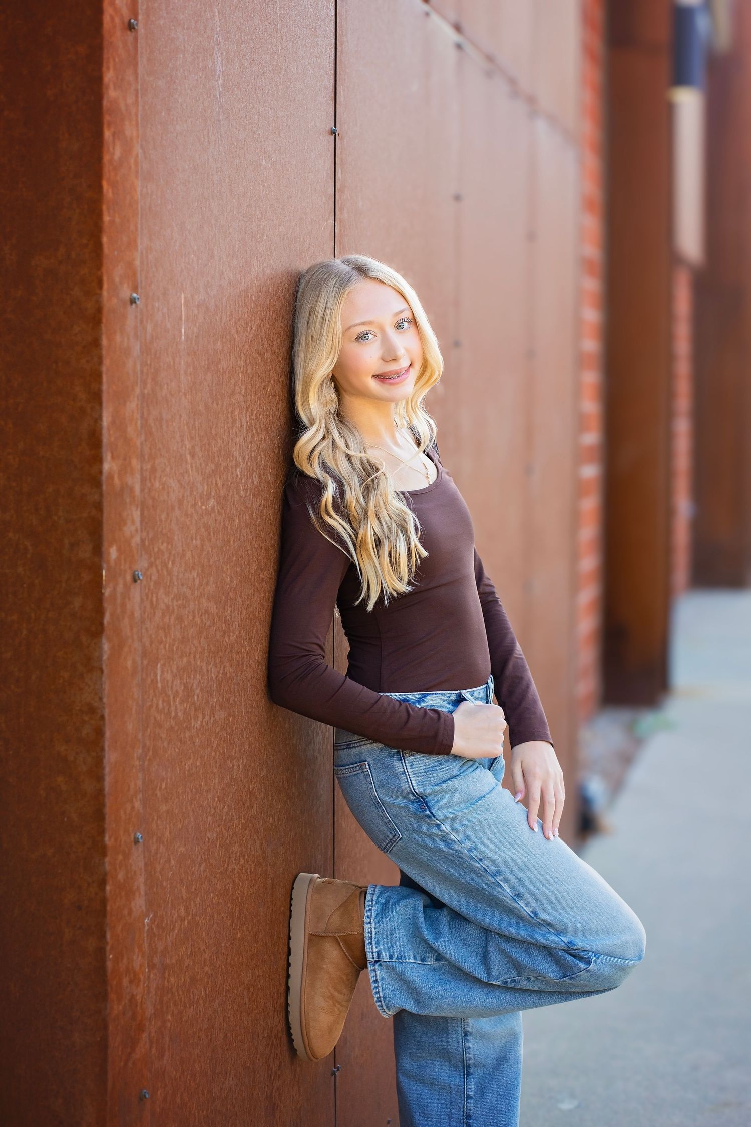 Blonde teen in brown top and jeans leaning against rusty metal wall outdoors.
