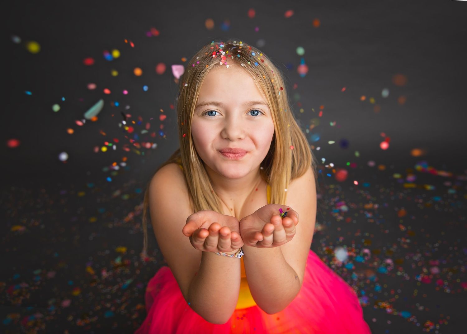 Young blonde girl in pink tutu blowing colorful confetti from her hands in a studio photo.