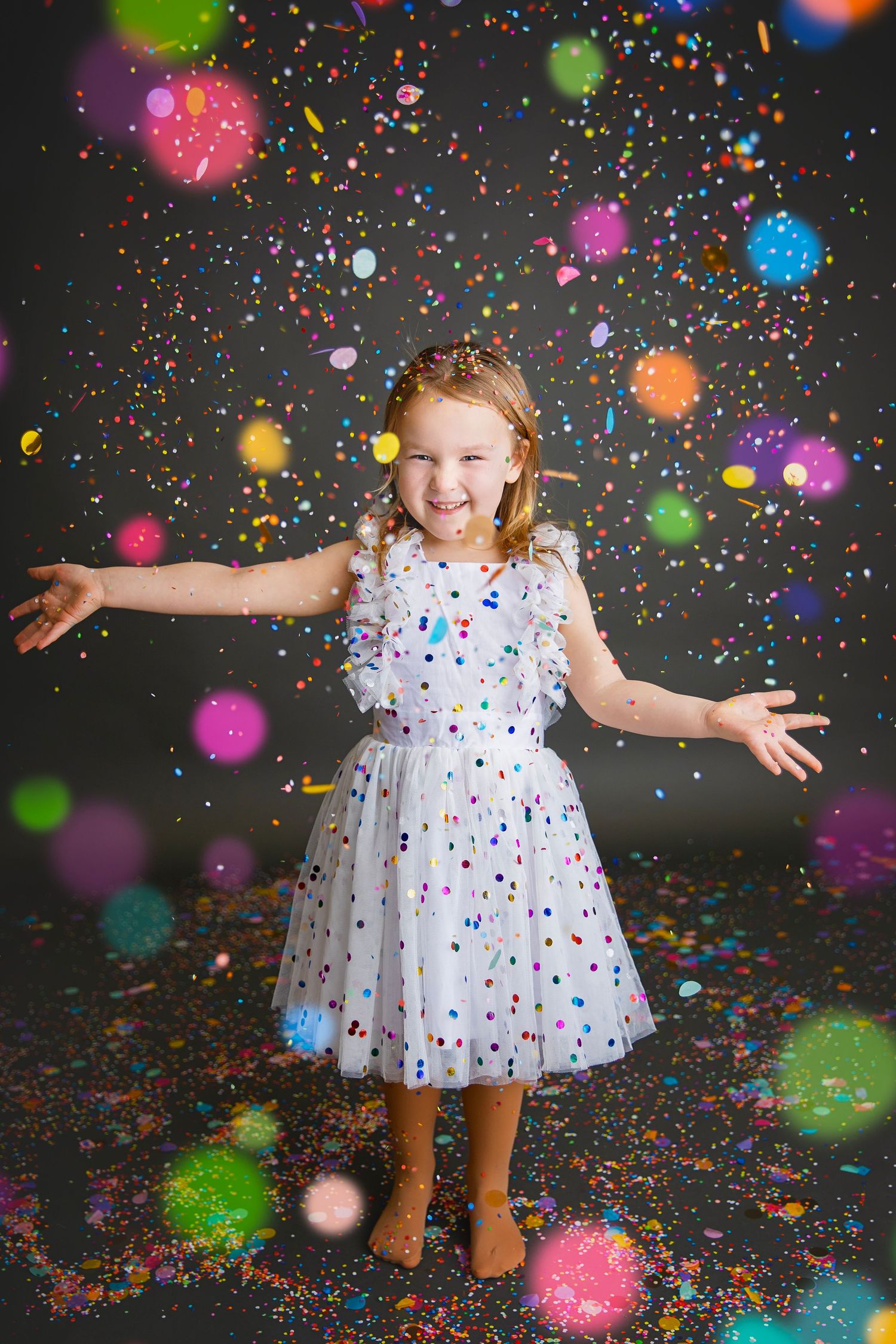 A laughing young girl in a white polka dot dress stands with arms outstretched amid falling colorful confetti.