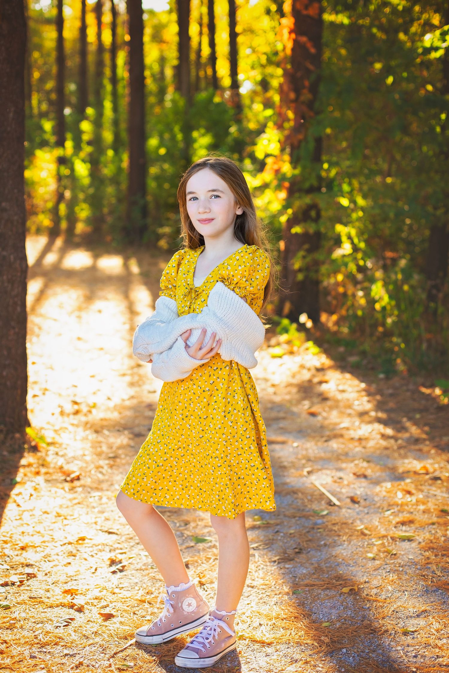 Young girl in a yellow floral dress standing on a sunlit autumn forest path.