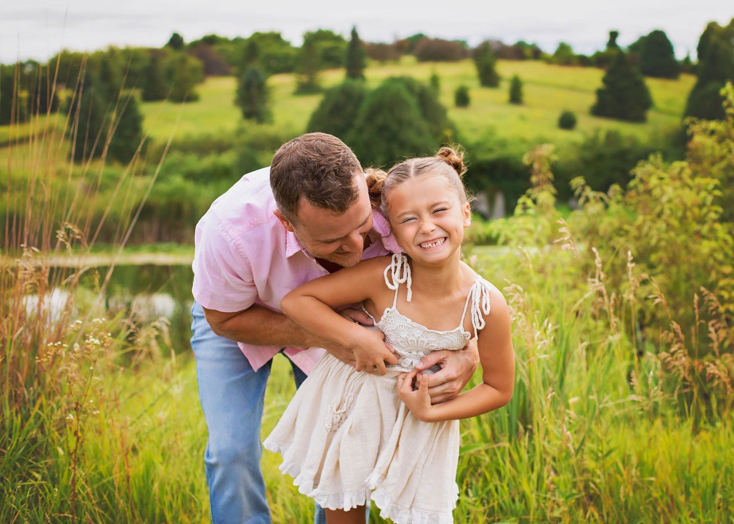 Father hugging laughing daughter in a lush green meadow during an outdoor family photo session.