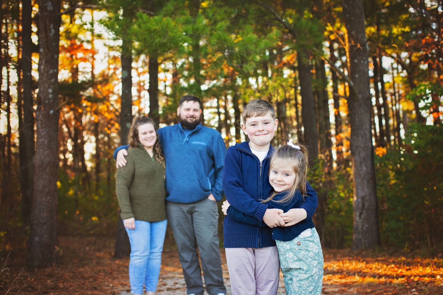 Family of four posing on woodland trail with two children in front and parents behind in fall foliage.