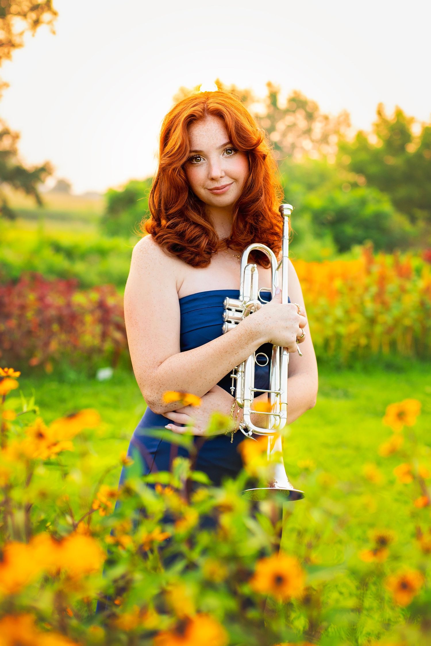 Young redhead woman holding a silver trumpet in a field of yellow wildflowers during golden hour.