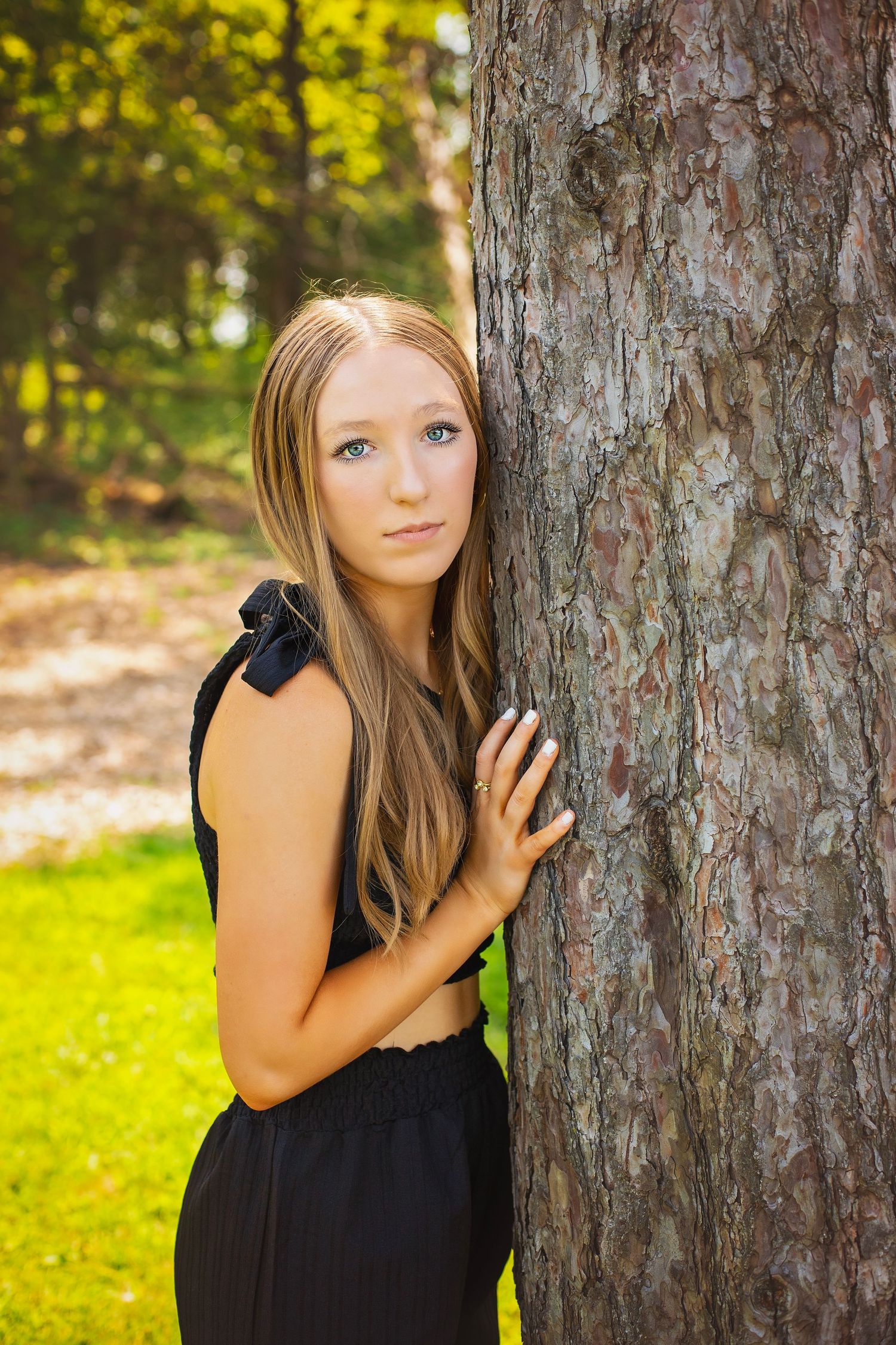 Teen girl with long blonde hair leans against a pine tree trunk in a sunny green park.