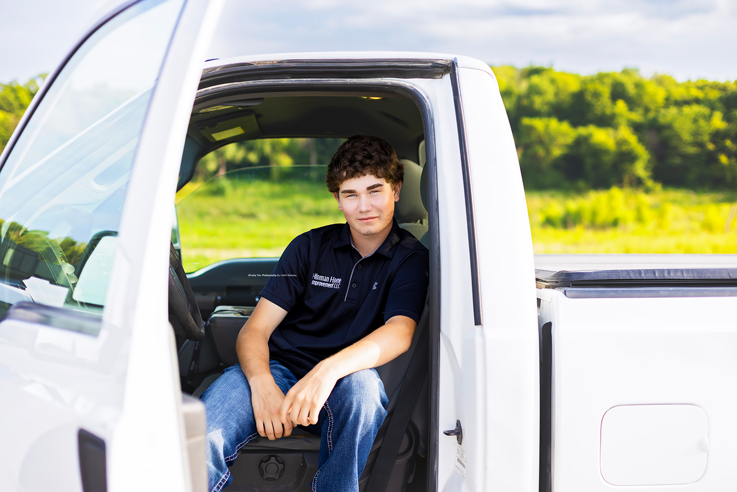 Young man in dark polo shirt sits in open door of white pickup truck with green fields in background.