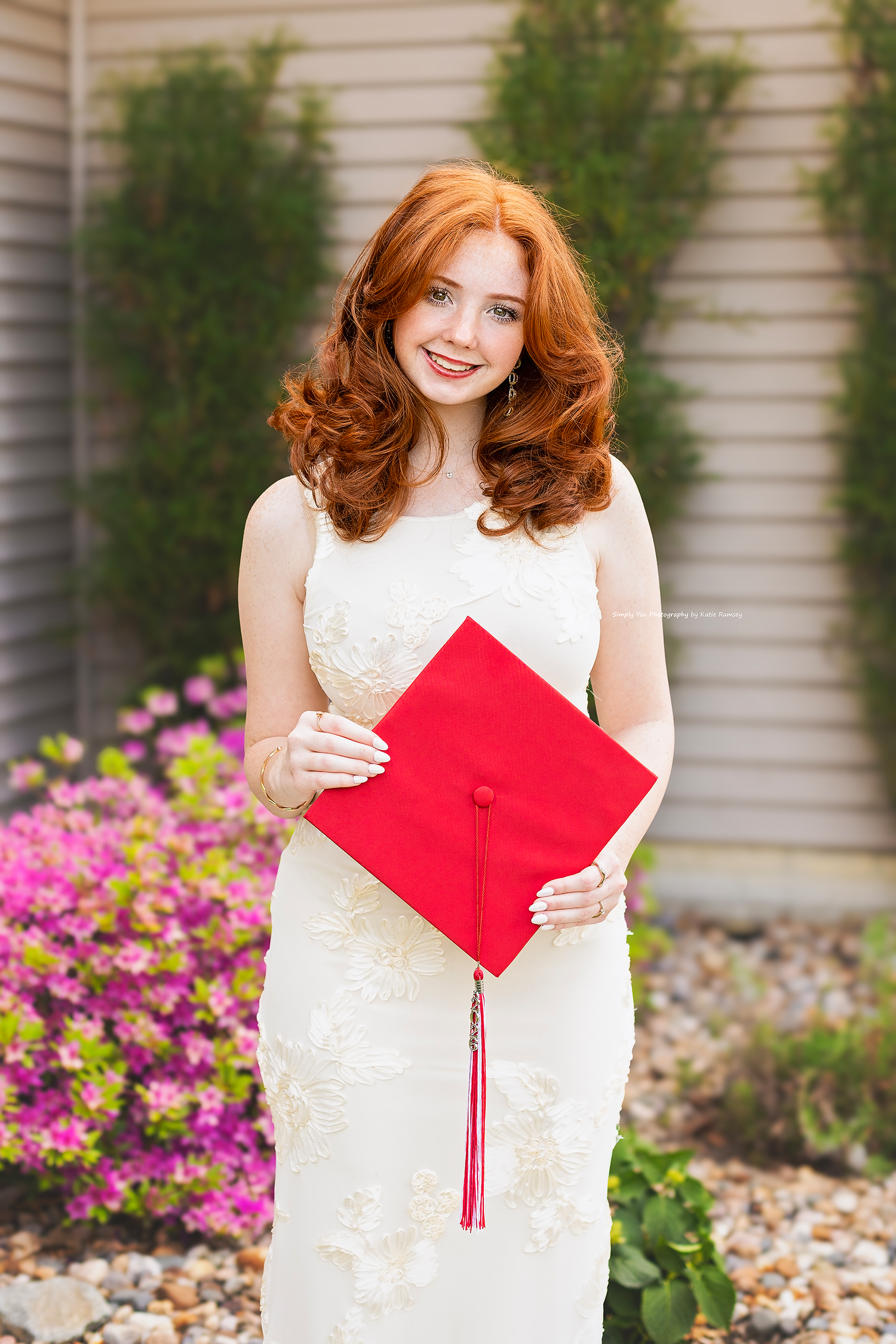 Smiling redhead graduate in white lace dress holding red mortarboard cap outdoors near pink flowers.