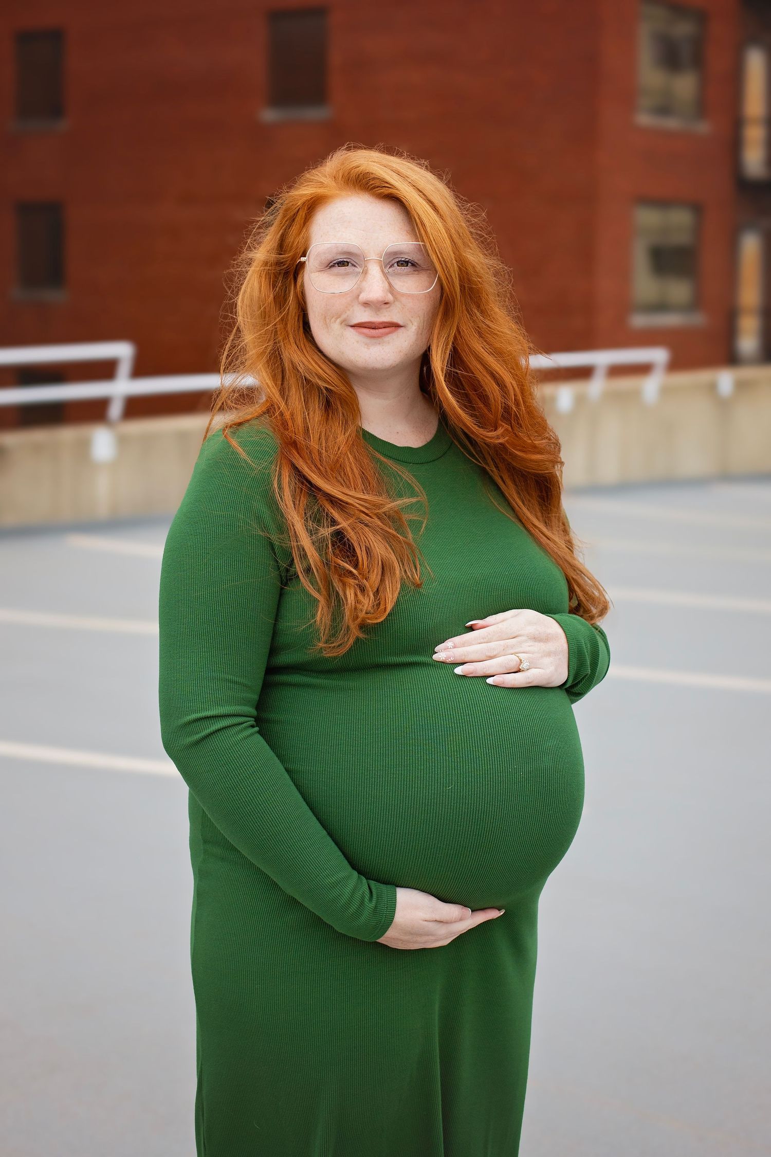 Pregnant woman in green dress with long red hair cradles her baby bump outdoors.